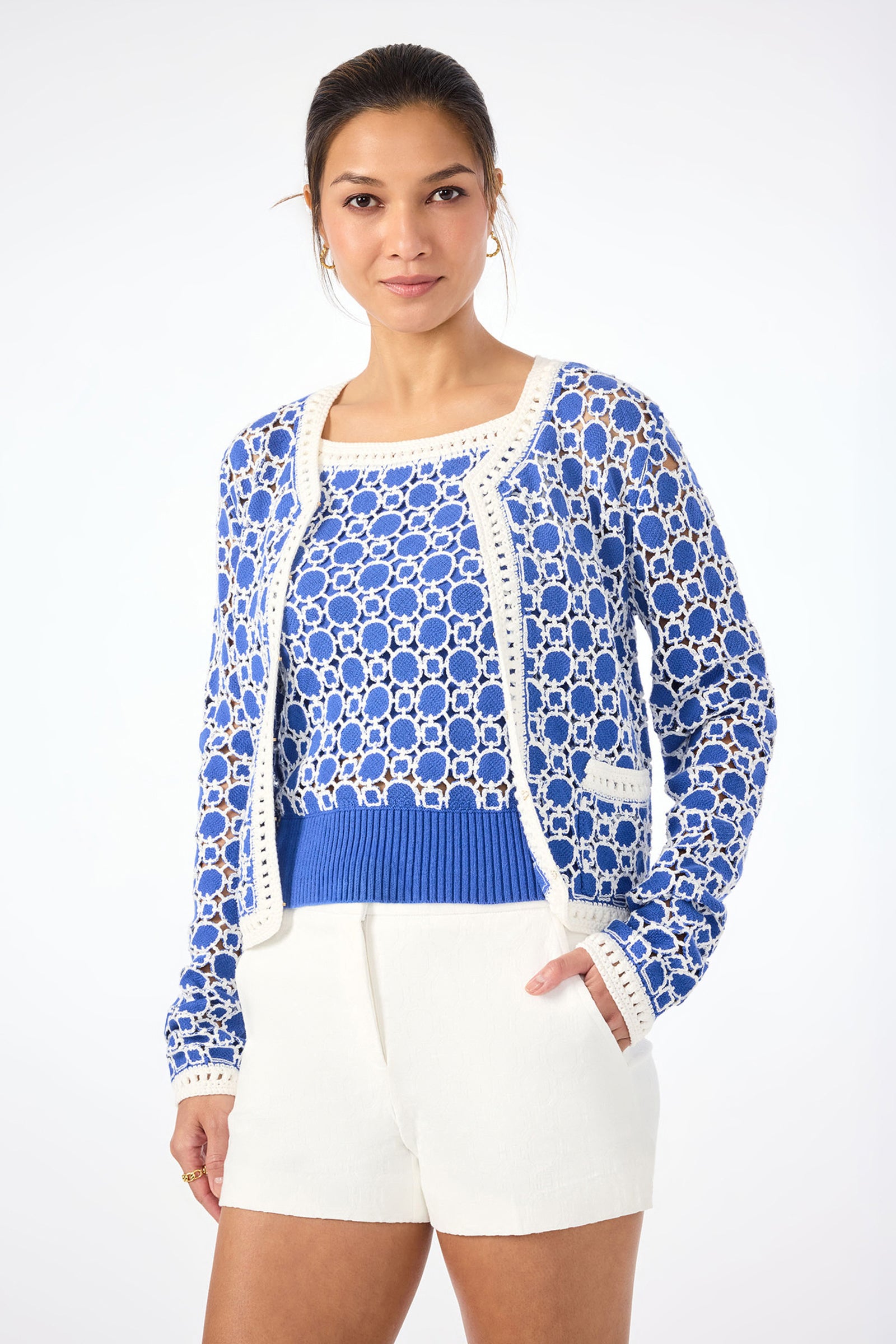 A woman stands against a plain white background, wearing the Sinai Contrast Trim Knit Cardigan over a matching blue top and white shorts. Her hair is tied back and her hands are in her pockets.