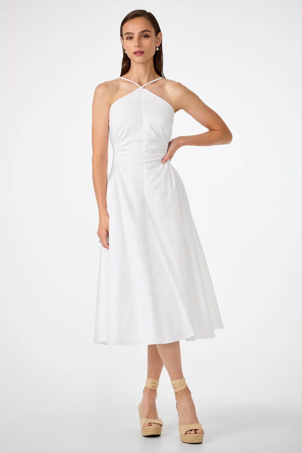 A woman with straight brown hair and a neutral expression wears the white Kata Shirred Waist Midi Dress, featuring a sleeveless, halter-neck design and flared skirt, paired with beige platform sandals against a white background.