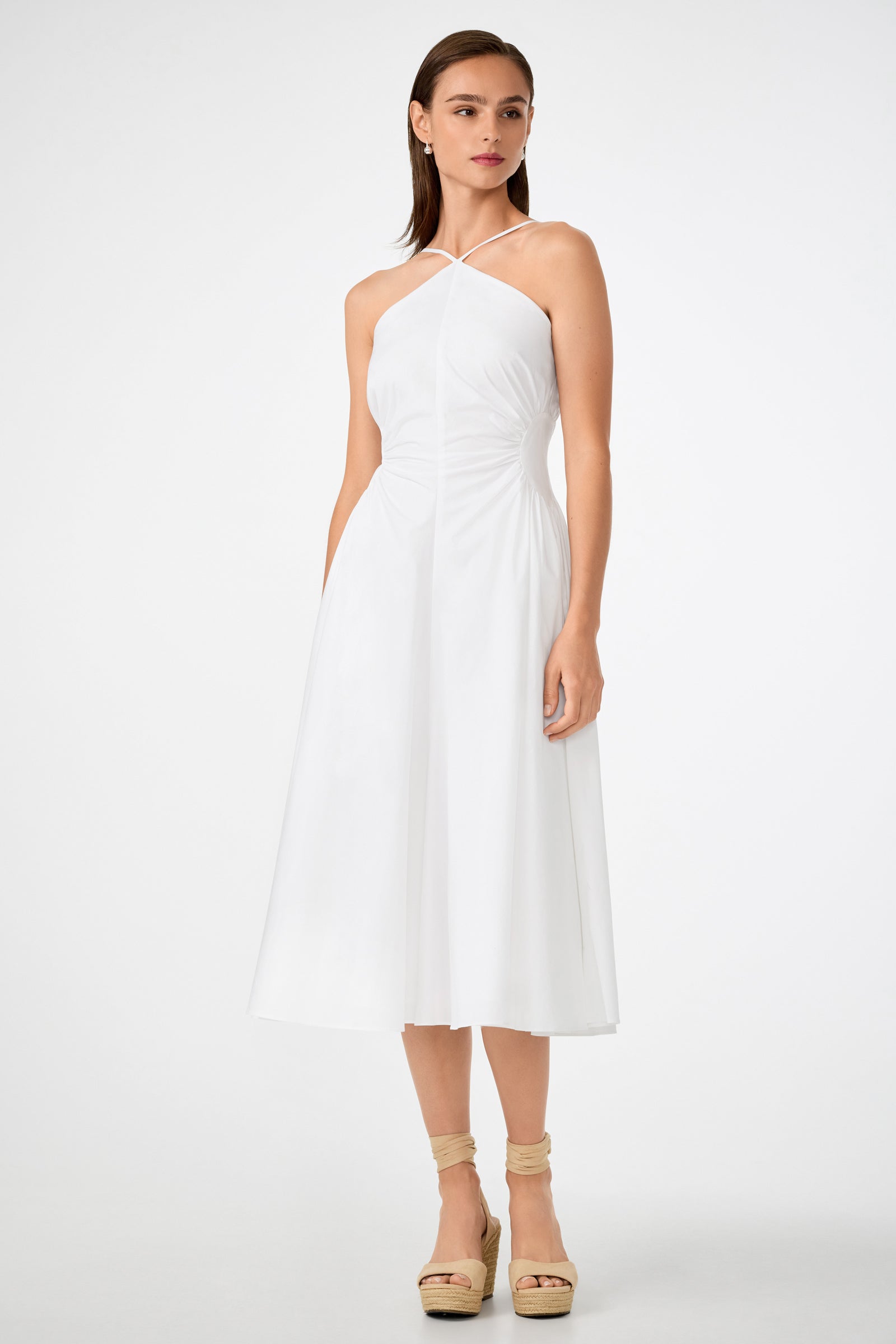 A woman with straight brown hair wears the Kata Shirred Waist Midi Dress, a sleeveless white halter-neck style with a fitted bodice and flared skirt. She pairs it with beige platform sandals and stands against a plain white background.