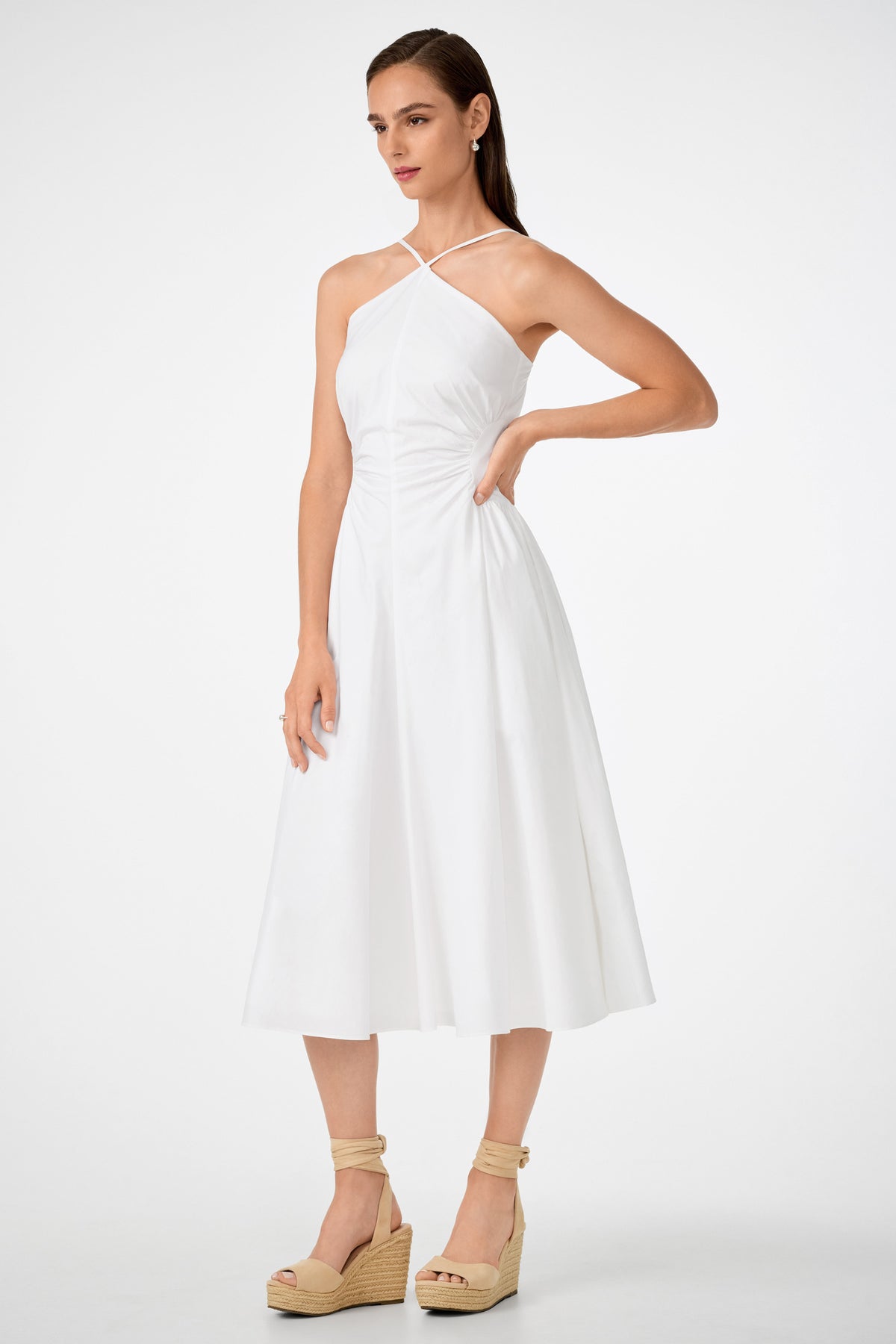 A woman wears the white Kata Shirred Waist Midi Dress with a halter neckline, paired with beige wedge sandals. She stands against a plain background, her long brown hair straight and one hand resting on her hip.