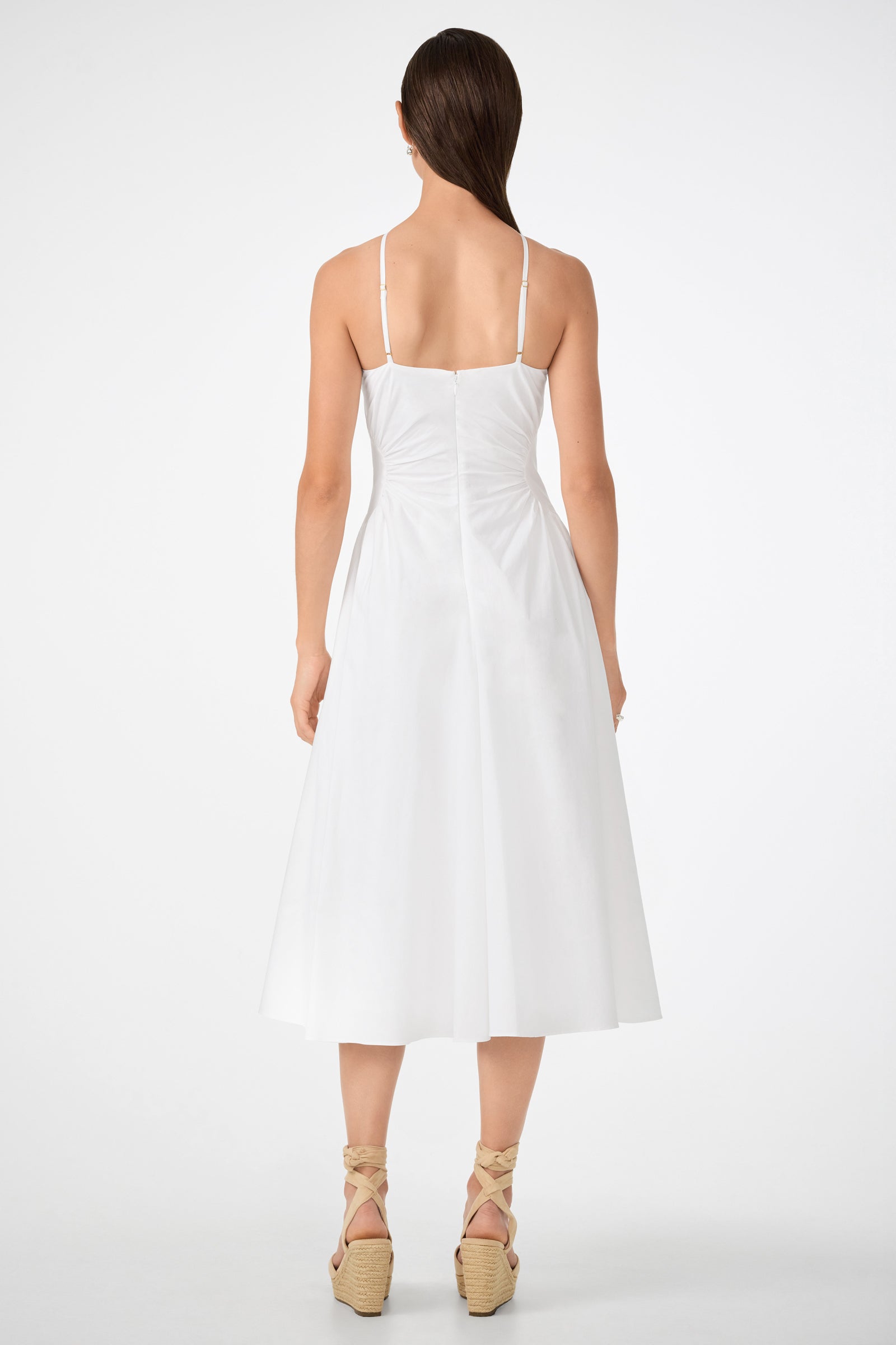 A woman with straight brown hair faces away, wearing the white Kata Shirred Waist Midi Dress—sleeveless, mid-length, with thin straps and a fitted bodice. She pairs it with wedge sandals featuring ankle ties against a plain white background.