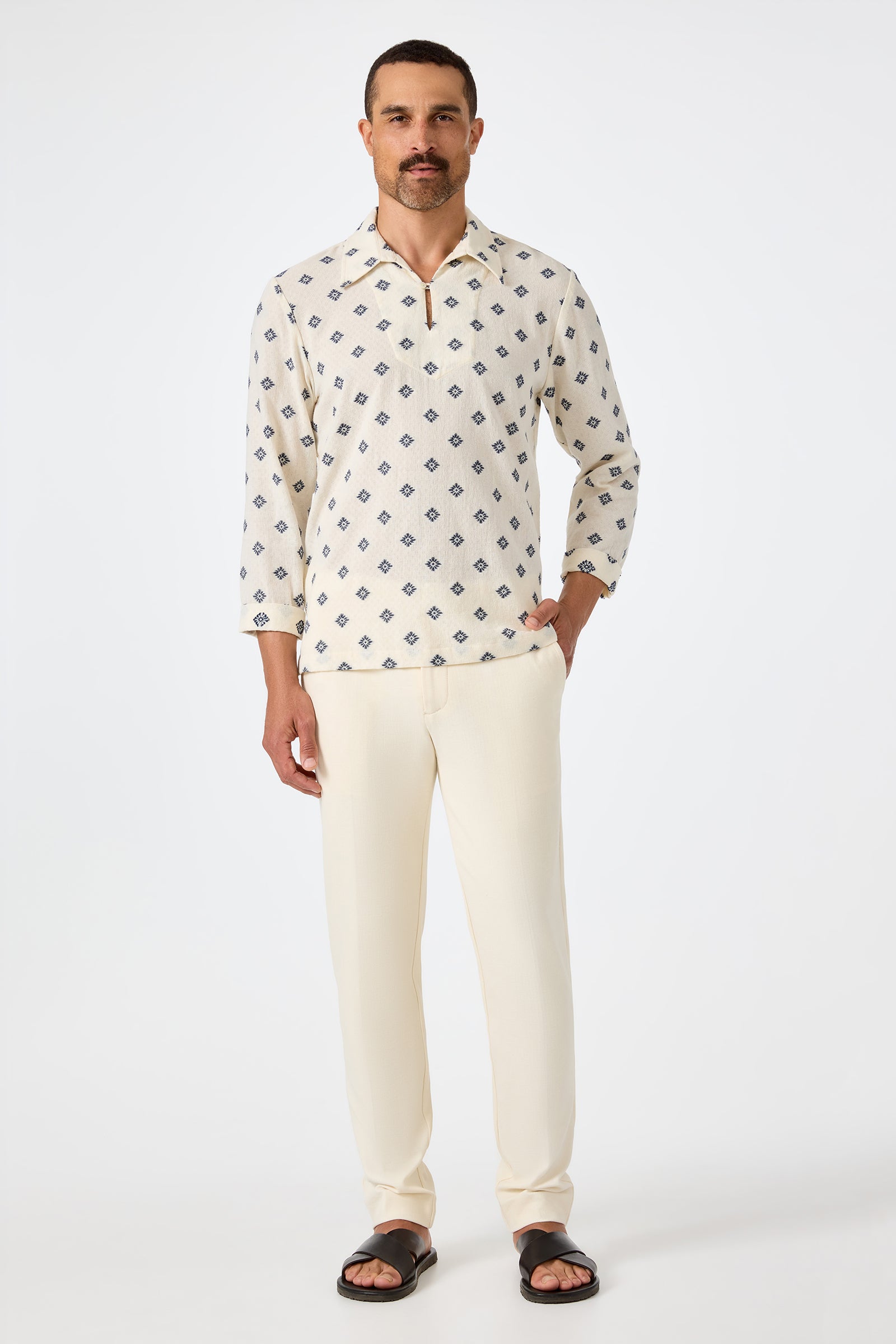 A man with short hair and a mustache stands against a white background, wearing the Hendrix Long-Sleeve Pullover in cream, cream pants, and black sandals. He poses with one hand in his pocket.