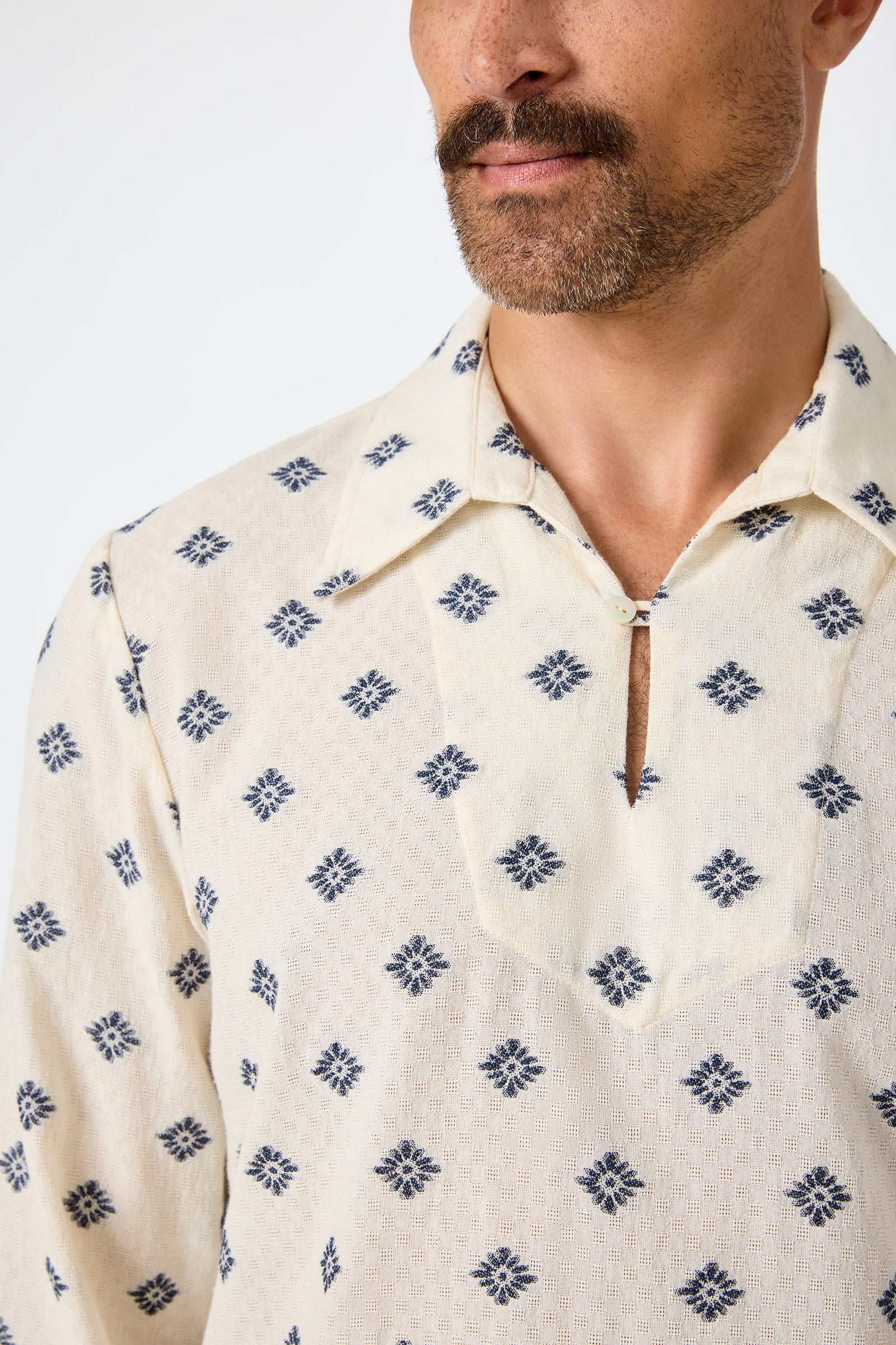 A man with a mustache is shown from the shoulders up against a plain background wearing the Hendrix Long-Sleeve Pullover—a cream-colored shirt with blue geometric print and a v-shaped notch at the collar.