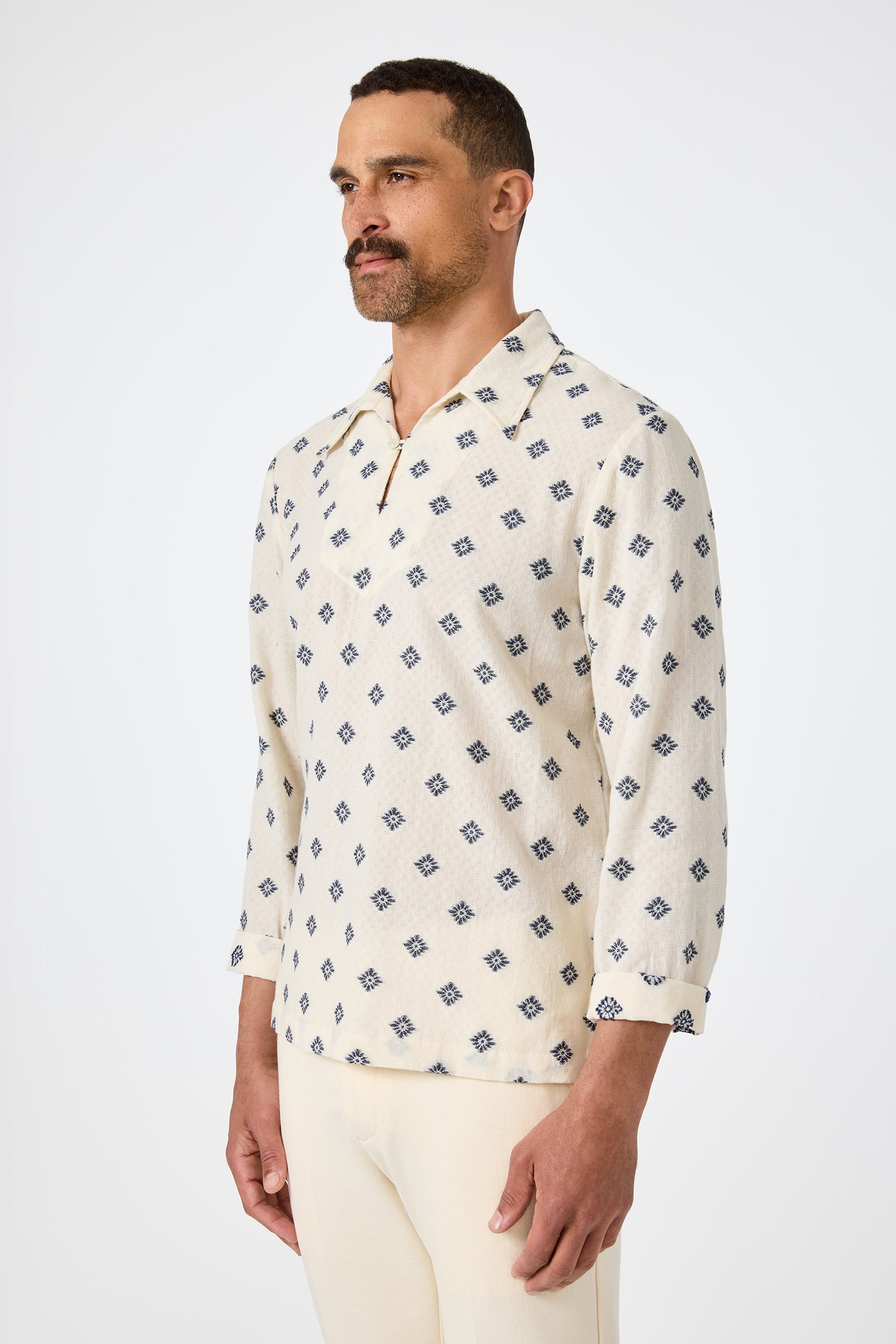 A man stands against a plain background, wearing the Hendrix Long-Sleeve Pullover—a cream shirt with a blue geometric pattern—and light pants. He faces slightly left with a neutral expression.