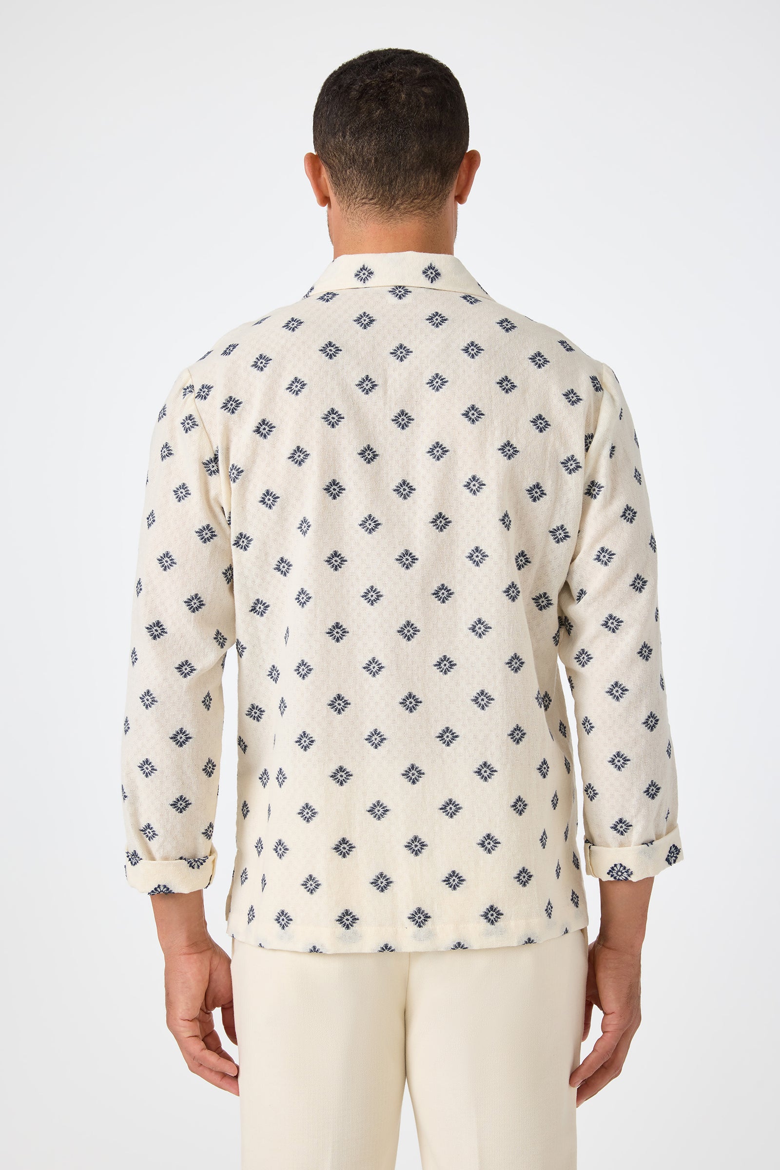 A man faces away from the camera, wearing the Hendrix Long-Sleeve Pullover in cream with a navy geometric diamond pattern and matching cream pants against a plain white background.