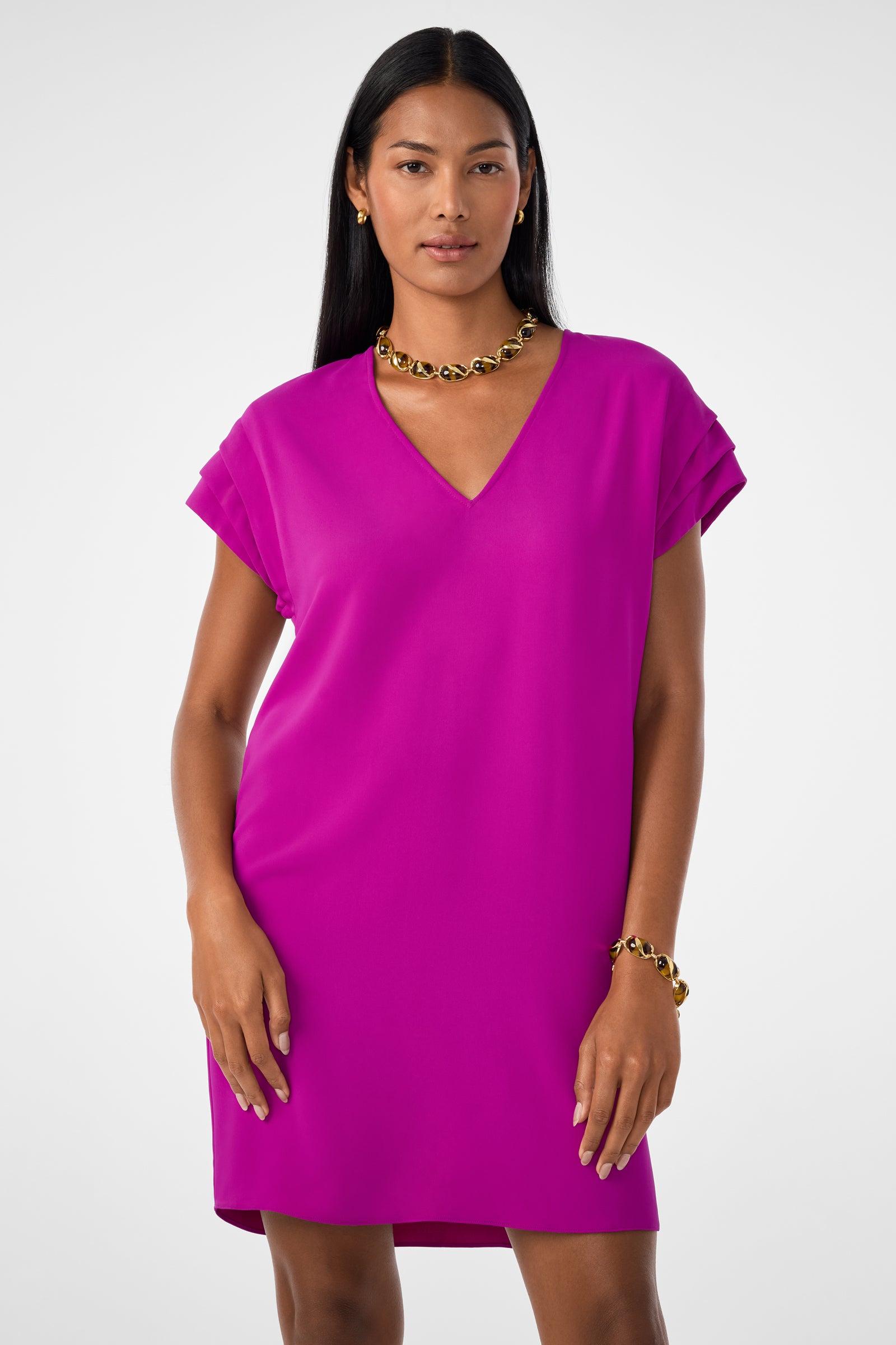 A woman with long straight hair, wearing a bright pink V-neck dress, stands facing forward against a plain light background. She accessorizes with a gold chain necklace and an Oval Tortoise Bracelet.