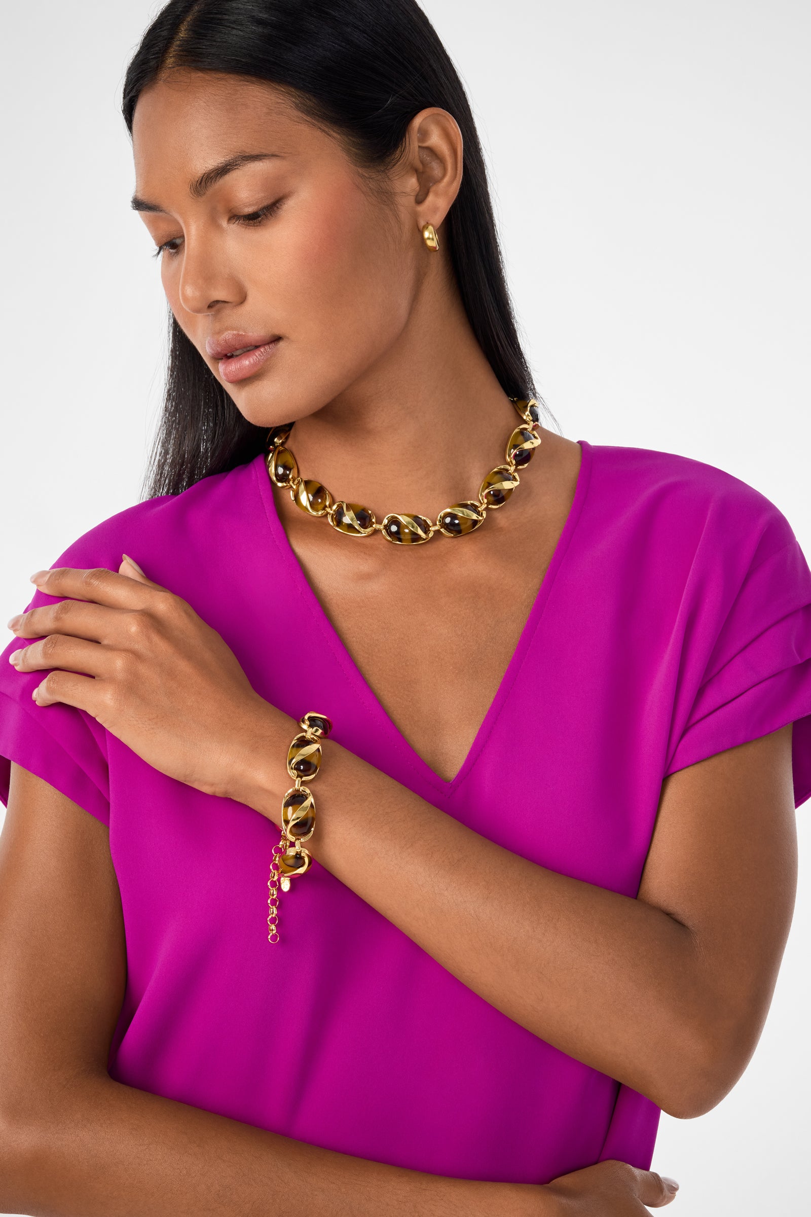 A woman in a bright magenta dress wears the Kjl Oval Tortoise Bracelet, paired with a chunky gold and black necklace and small gold earrings. She looks down with her arms gently crossed over her chest against a plain background.