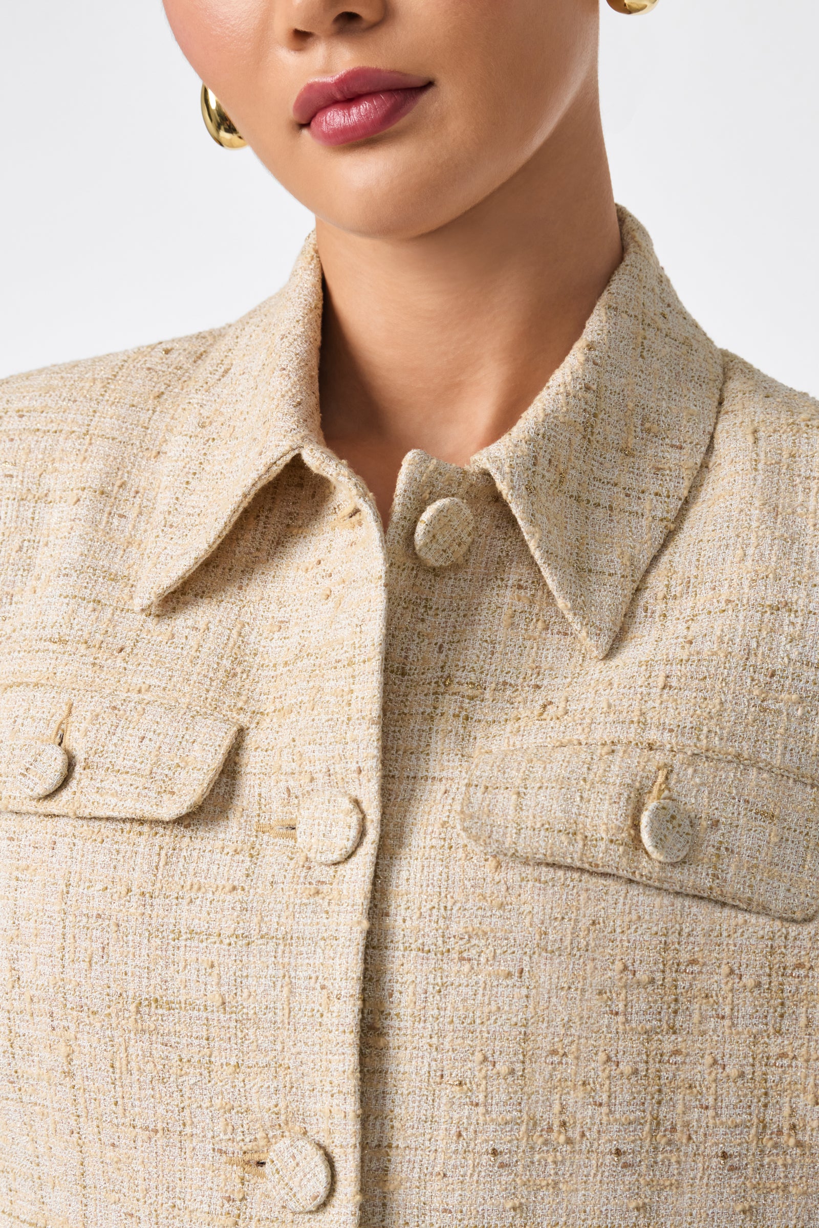 A woman shown from the shoulders up against a plain background wears the Bayonne Jacket, a beige textured button-up with flap pockets, paired with gold hoop earrings.