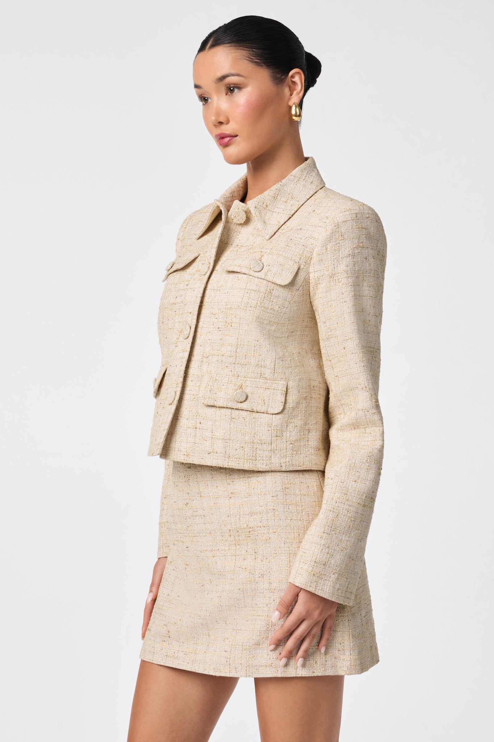 A woman stands against a plain white background wearing the Bayonne Jacket in beige—a textured, long-sleeve piece with four front pockets—paired with a matching mini skirt.