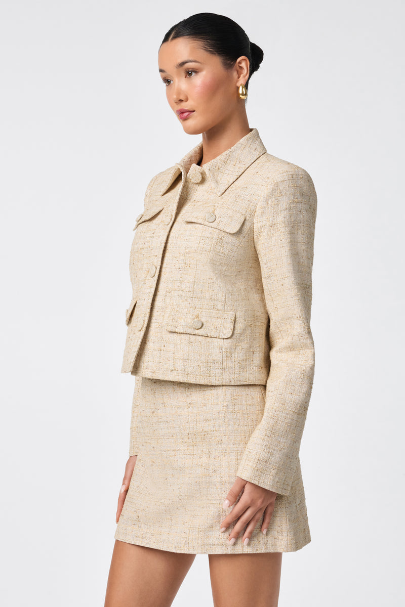 A woman stands against a plain white background wearing the Bayonne Jacket in beige—a textured, long-sleeve piece with four front pockets—paired with a matching mini skirt.