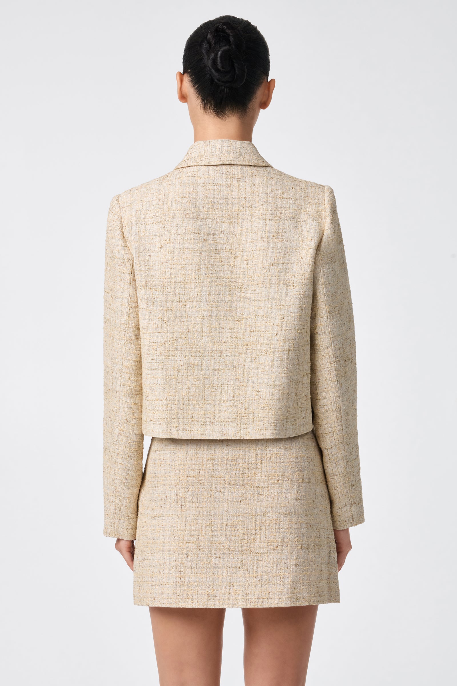 A woman stands with her back to the camera, wearing the Bayonne Jacket in beige with a matching skirt. Her hair is in a neat bun against a plain white background.