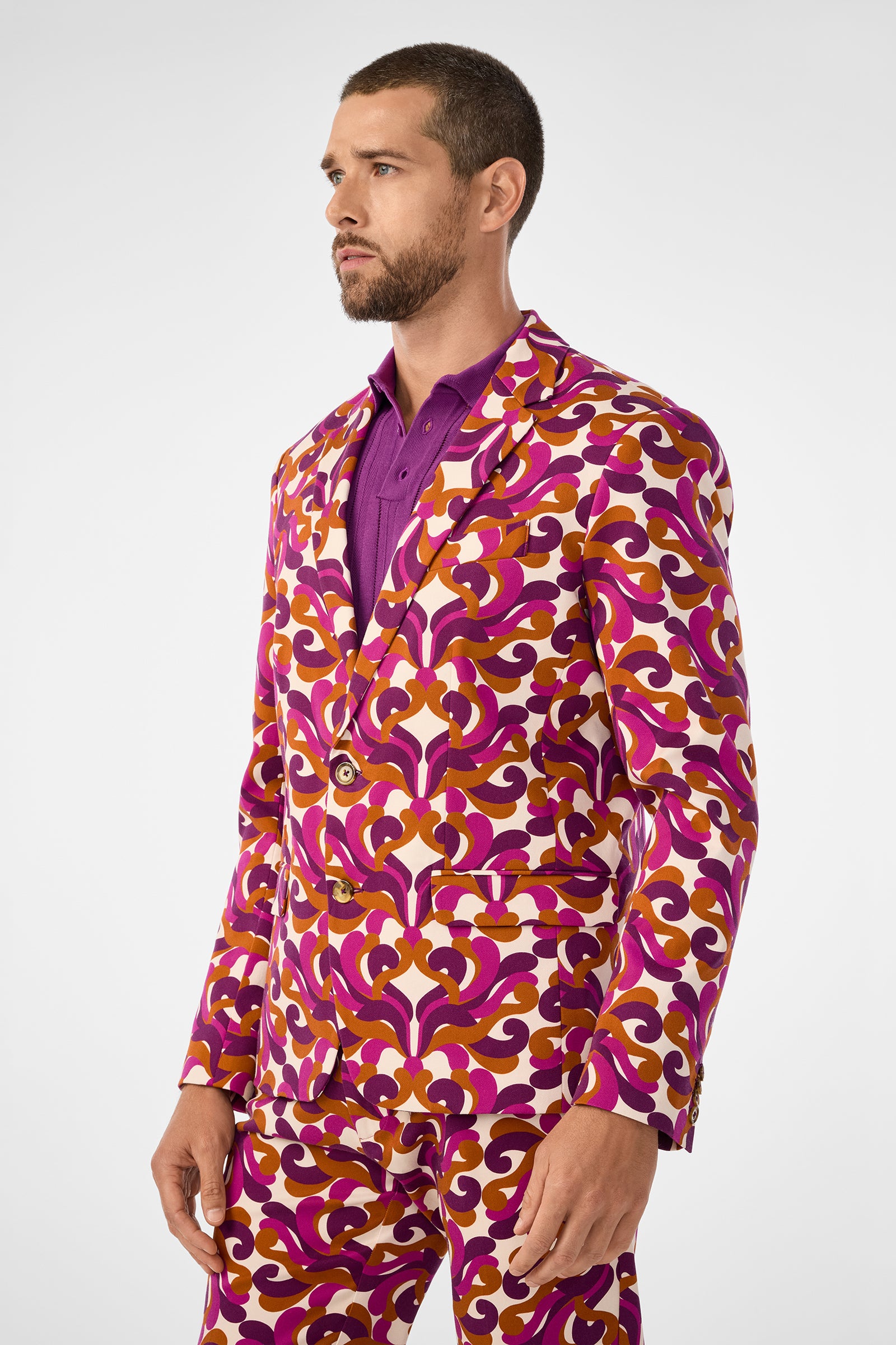 A man with short hair and a beard wears the Thurston Blazer—a colorful patterned suit in purple, pink, orange, and white—over a purple shirt, standing against a plain light background.