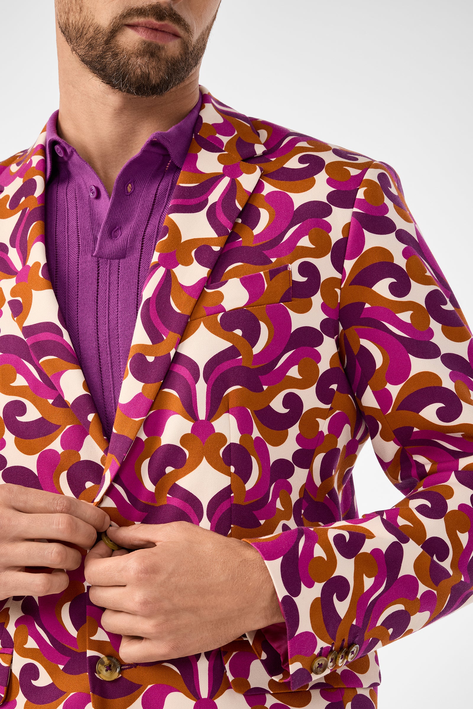 A man adjusts the button on his Thurston Blazer, which features a bold retro pattern in purple, pink, and orange over a purple shirt. The image is cropped to show his torso and part of his face.
