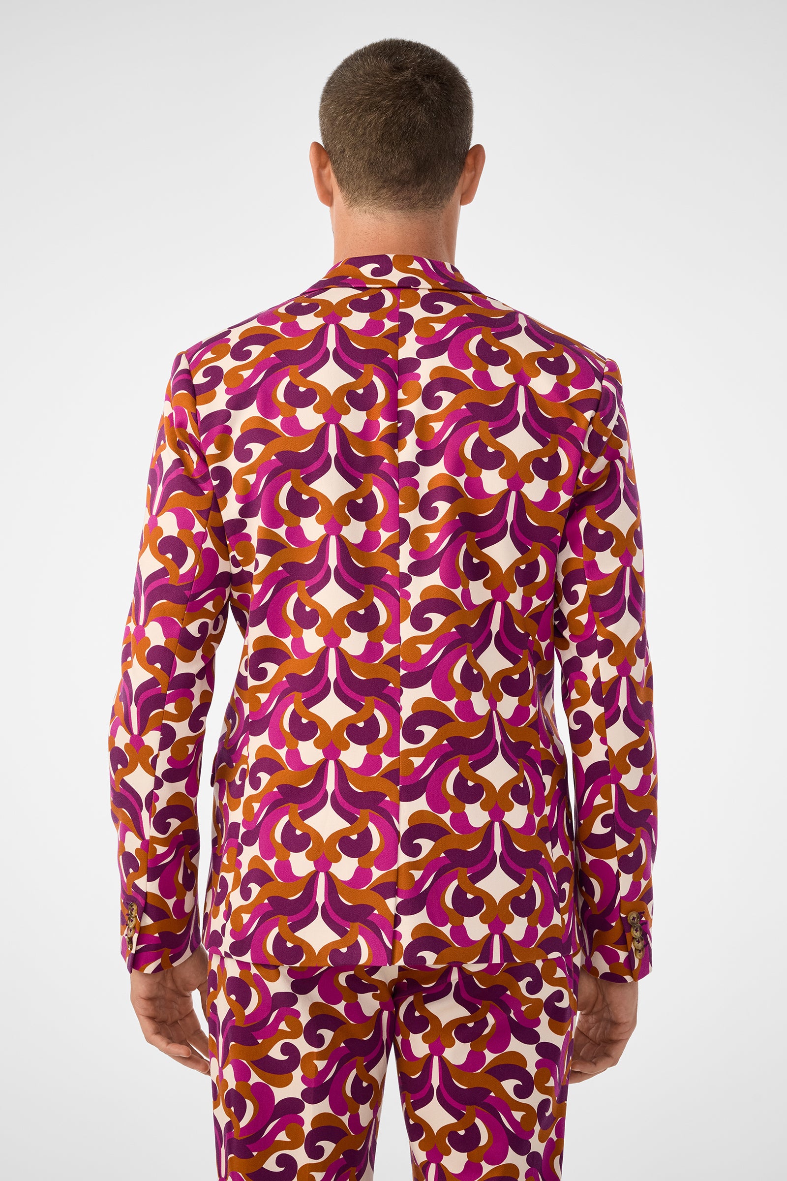 A man stands facing away, wearing the Thurston Blazer with matching trousers. Both feature a bold retro pattern in pink, purple, orange, and white with an intricate geometric design.