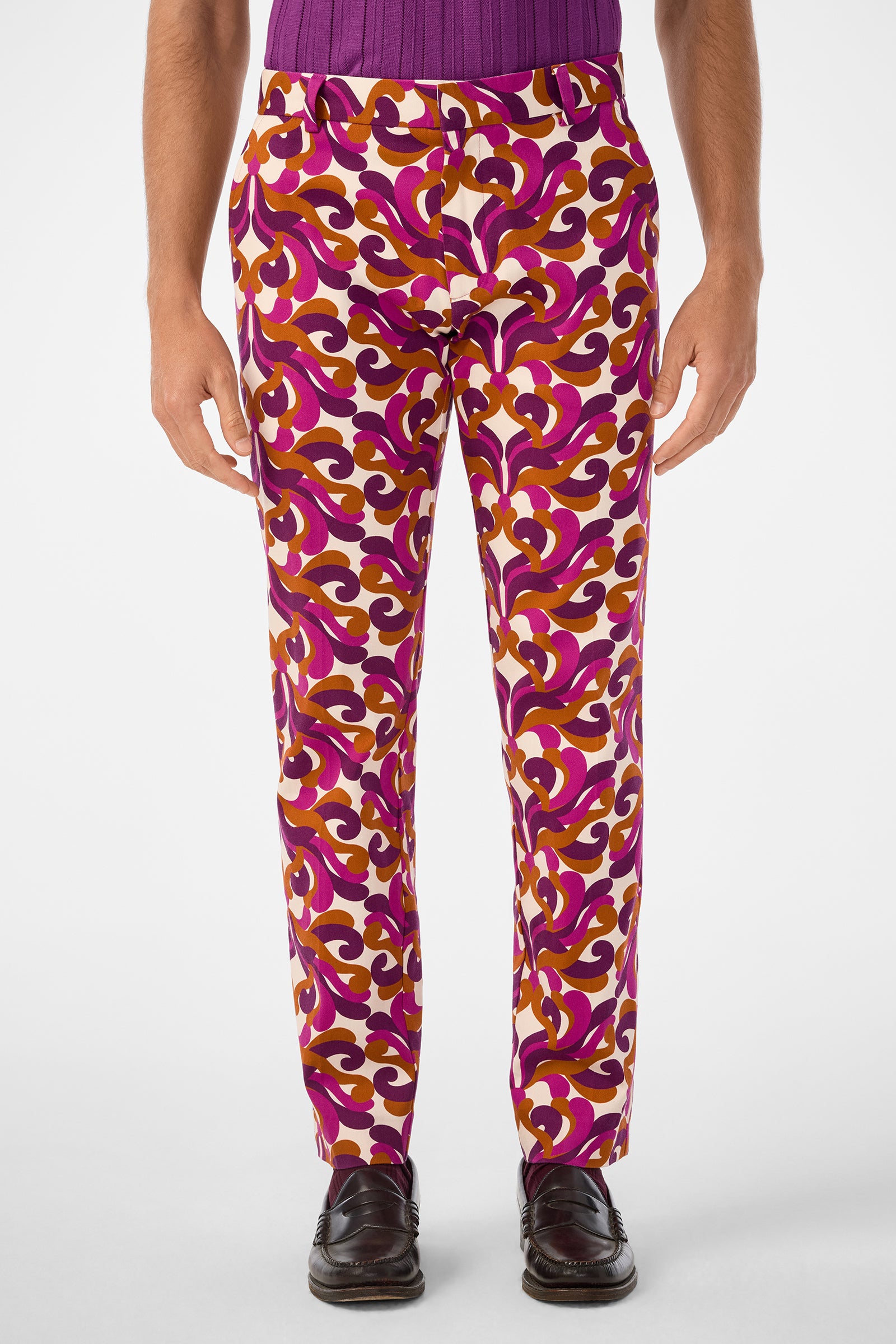 Someone wearing the Clyde Slim Trouser, featuring vibrant swirling purple, pink, and gold patterns, paired with a purple shirt and dark dress shoes, stands against a plain white background.