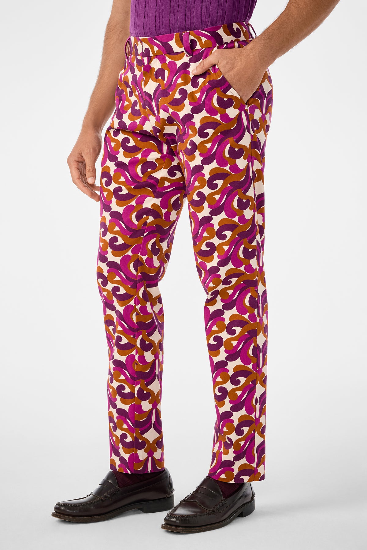 A person wearing Clyde Slim Trouser with swirling pink, purple, and yellow patterns, paired with a purple shirt and dark loafers, stands against a plain background with one hand in their pocket.