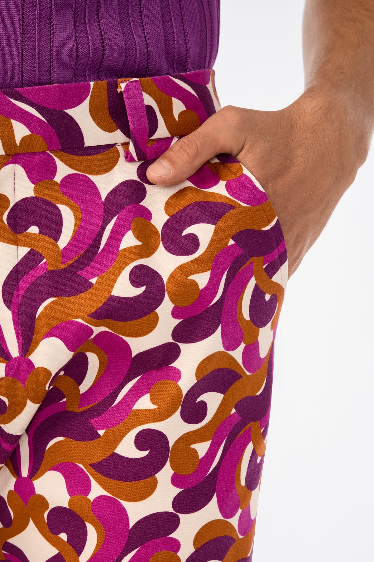 A person wears the Clyde Slim Trouser, featuring a bold retro pattern in pink, purple, orange, and brown, paired with a purple top. Their hand rests casually in the trouser pocket.