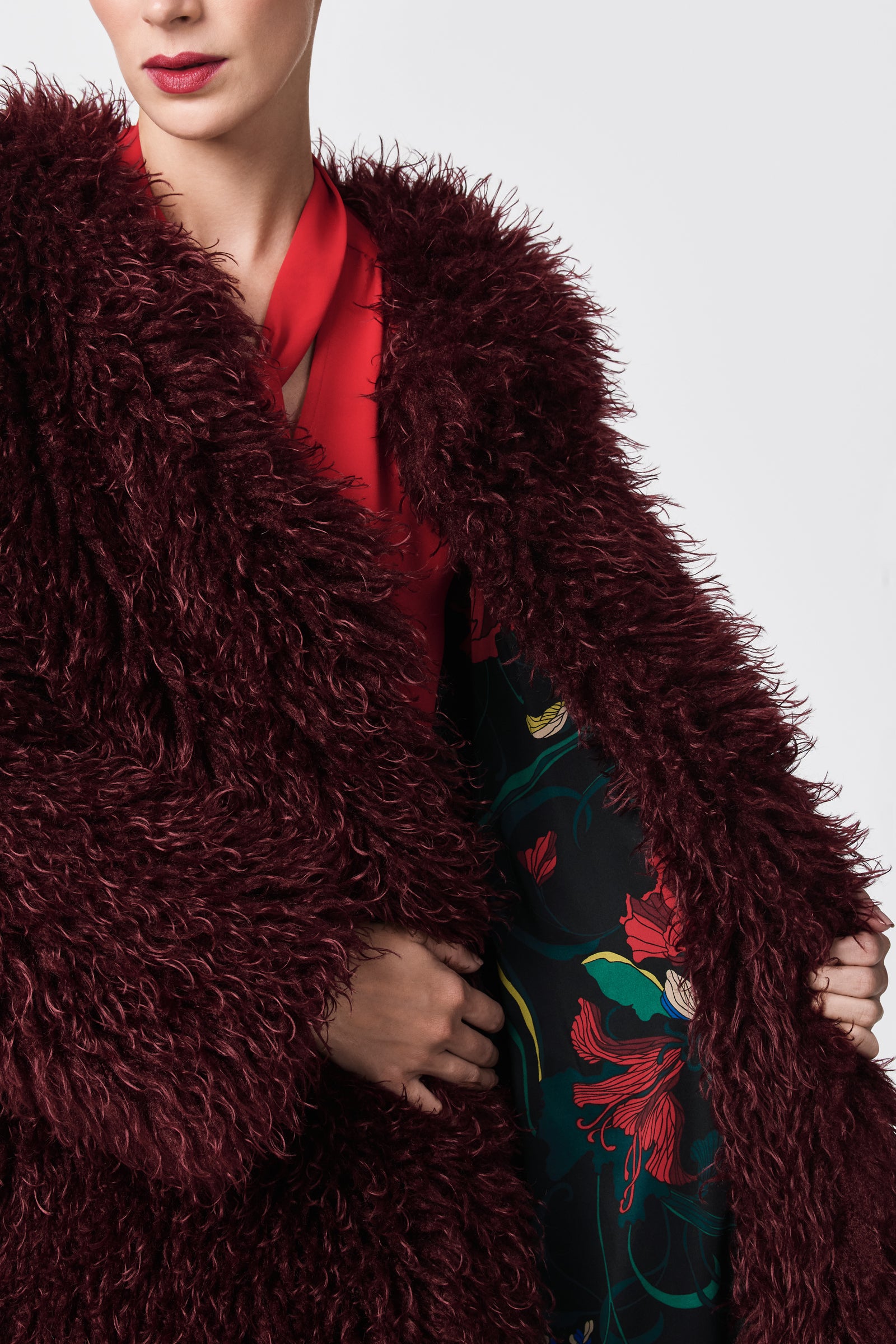 A person wears the Gladis Faux Fur Coat in deep burgundy over a red top, its open front revealing a black lining with red and green floral patterns. The background is plain light gray.