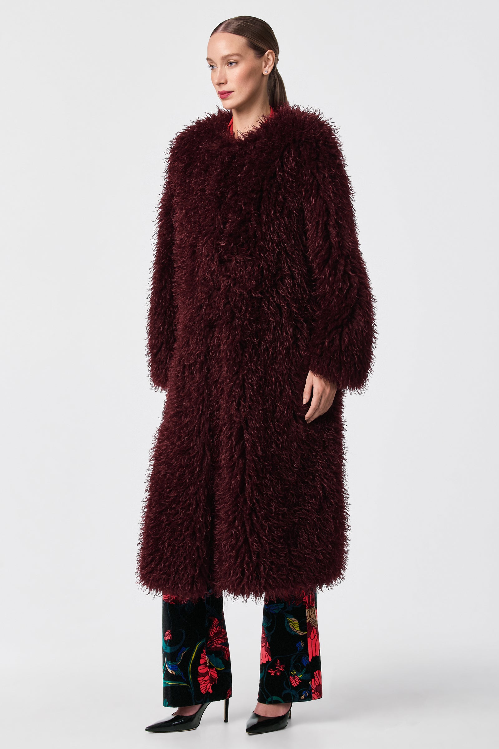 A woman stands against a plain background wearing the Gladis Faux Fur Coat in burgundy over floral pants and black heels. Her hair is tied back, and she gazes slightly to the side.