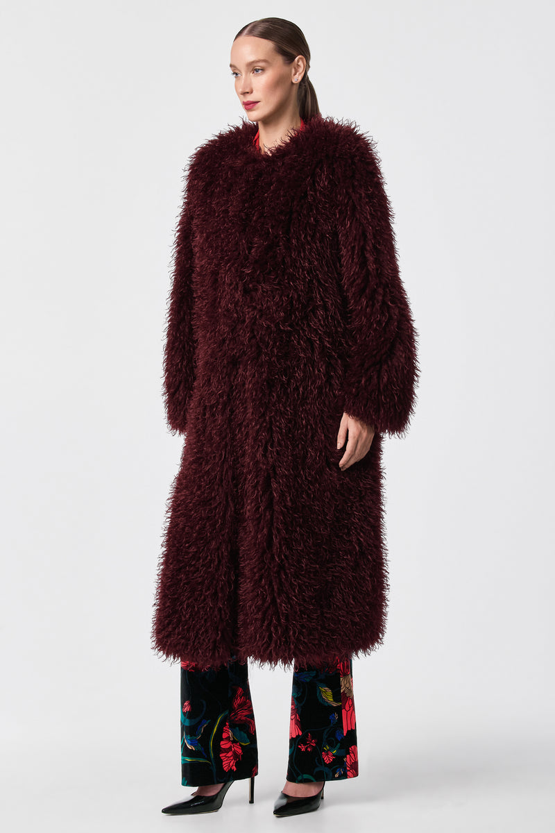 A woman stands against a plain background wearing the Gladis Faux Fur Coat in burgundy over floral pants and black heels. Her hair is tied back, and she gazes slightly to the side.