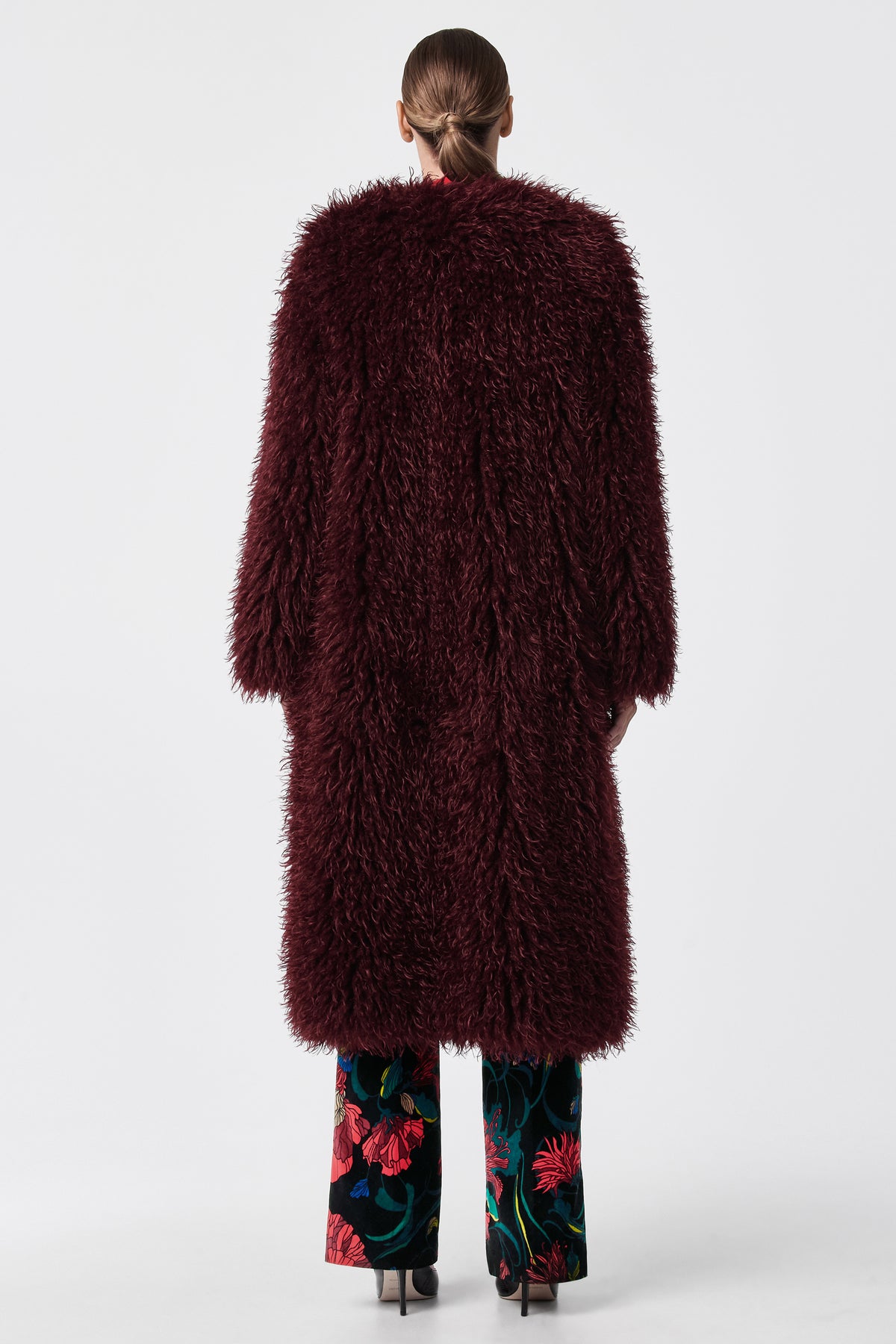 A person stands with their back to the camera, wearing the Gladis Faux Fur Coat in dark burgundy over floral-patterned trousers and black heels, set against a plain white background.