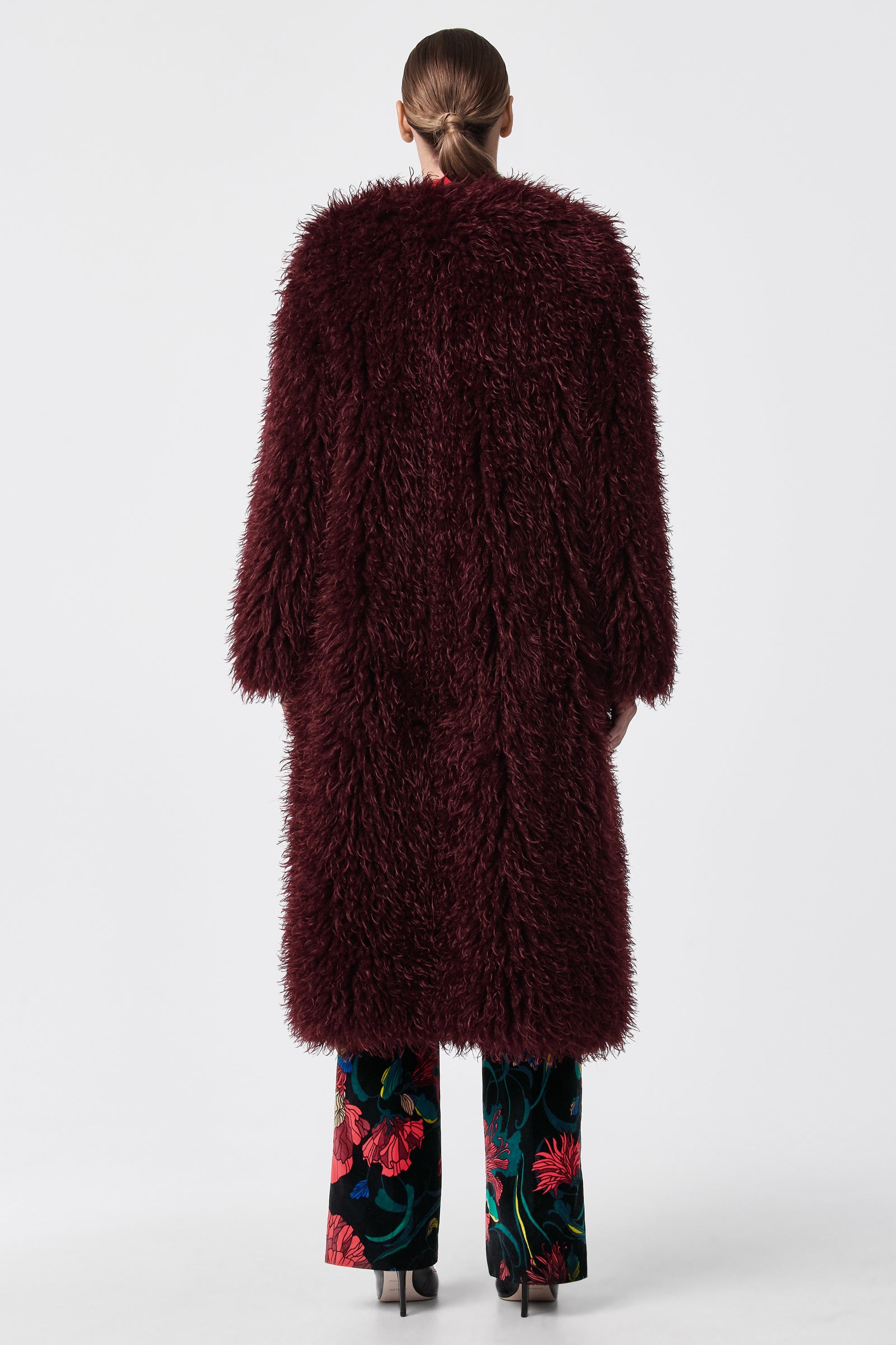 A person stands with their back to the camera, wearing the Gladis Faux Fur Coat in dark burgundy over floral-patterned trousers and black heels, set against a plain white background.