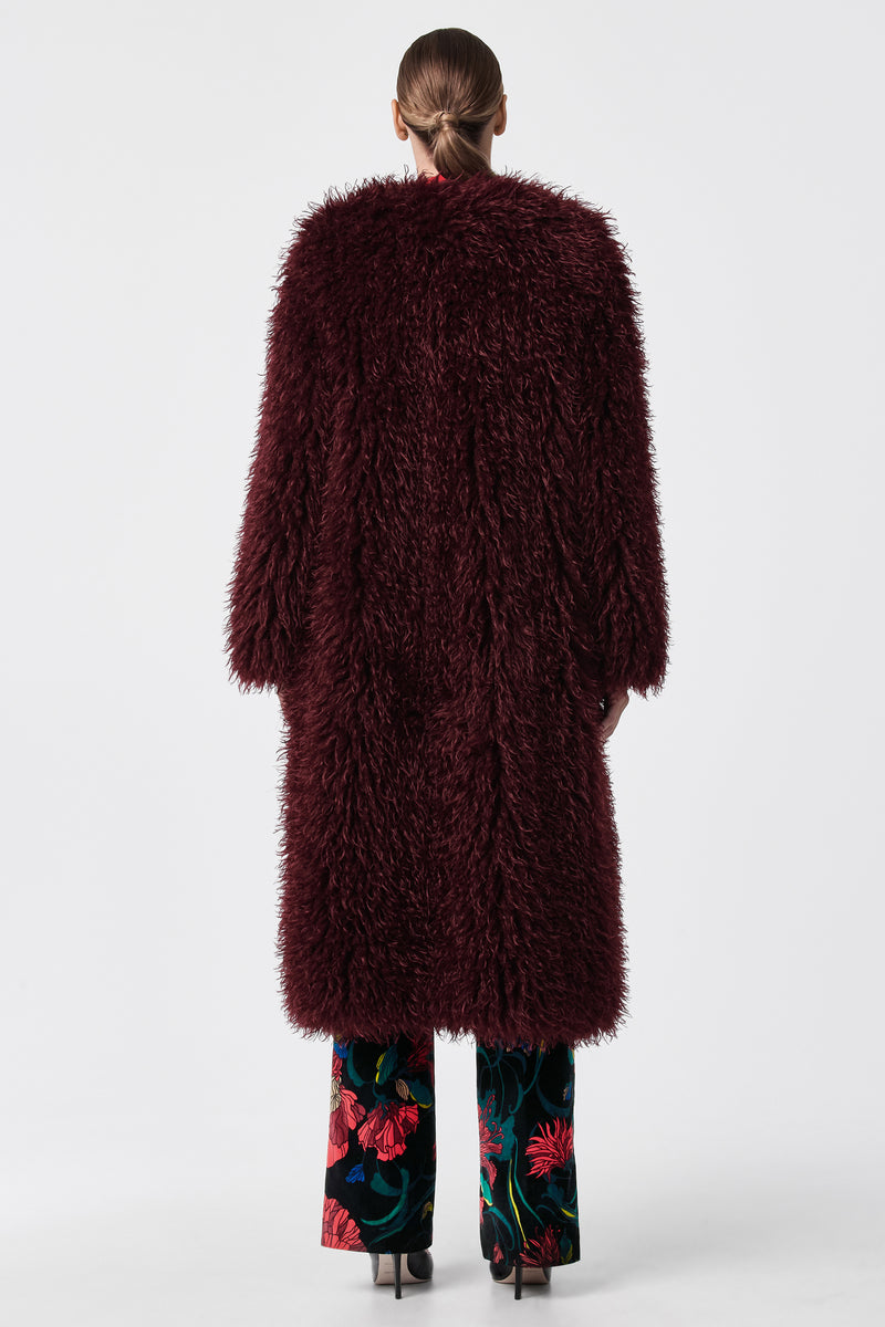 A person stands with their back to the camera, wearing the Gladis Faux Fur Coat in dark burgundy over floral-patterned trousers and black heels, set against a plain white background.