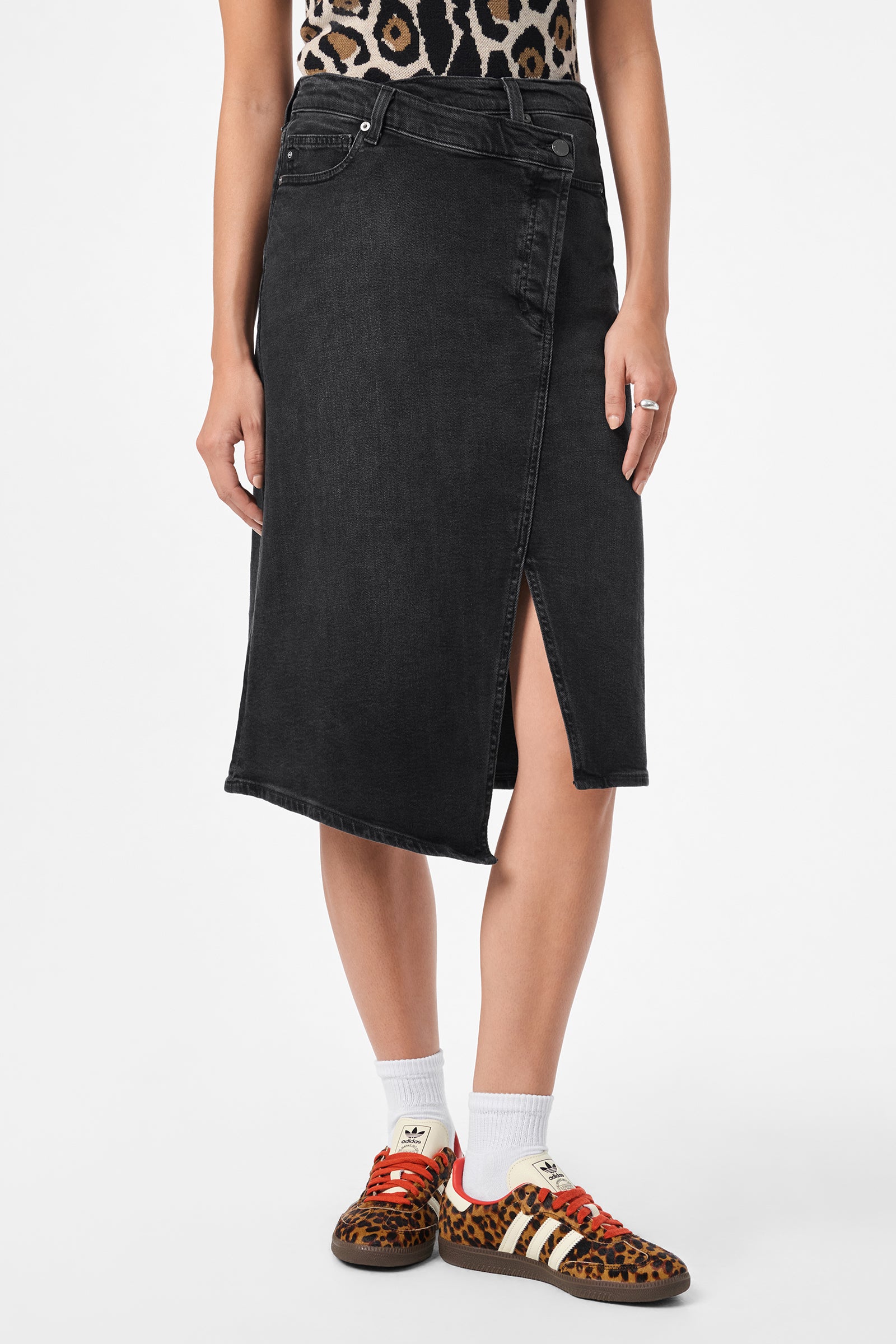 A person wearing the AG Bruna Skirt, an asymmetrical black denim skirt with a front slit, paired with a leopard print top, white socks, and brown leopard print Adidas sneakers, stands against a plain white background.