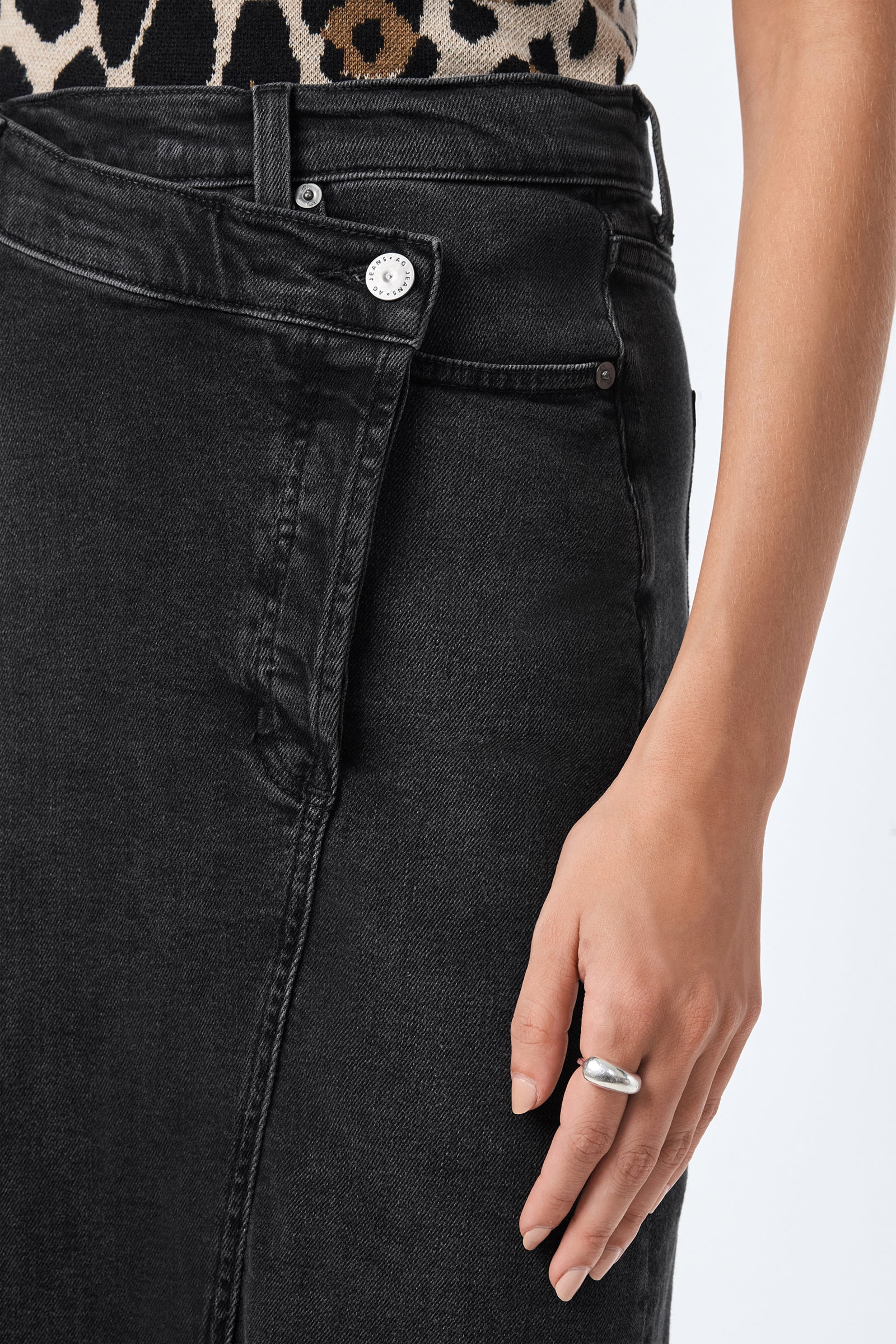 A close-up of a person wearing the AG Bruna Skirt in black denim, paired with a leopard print top and a silver ring on their right hand resting by their side.