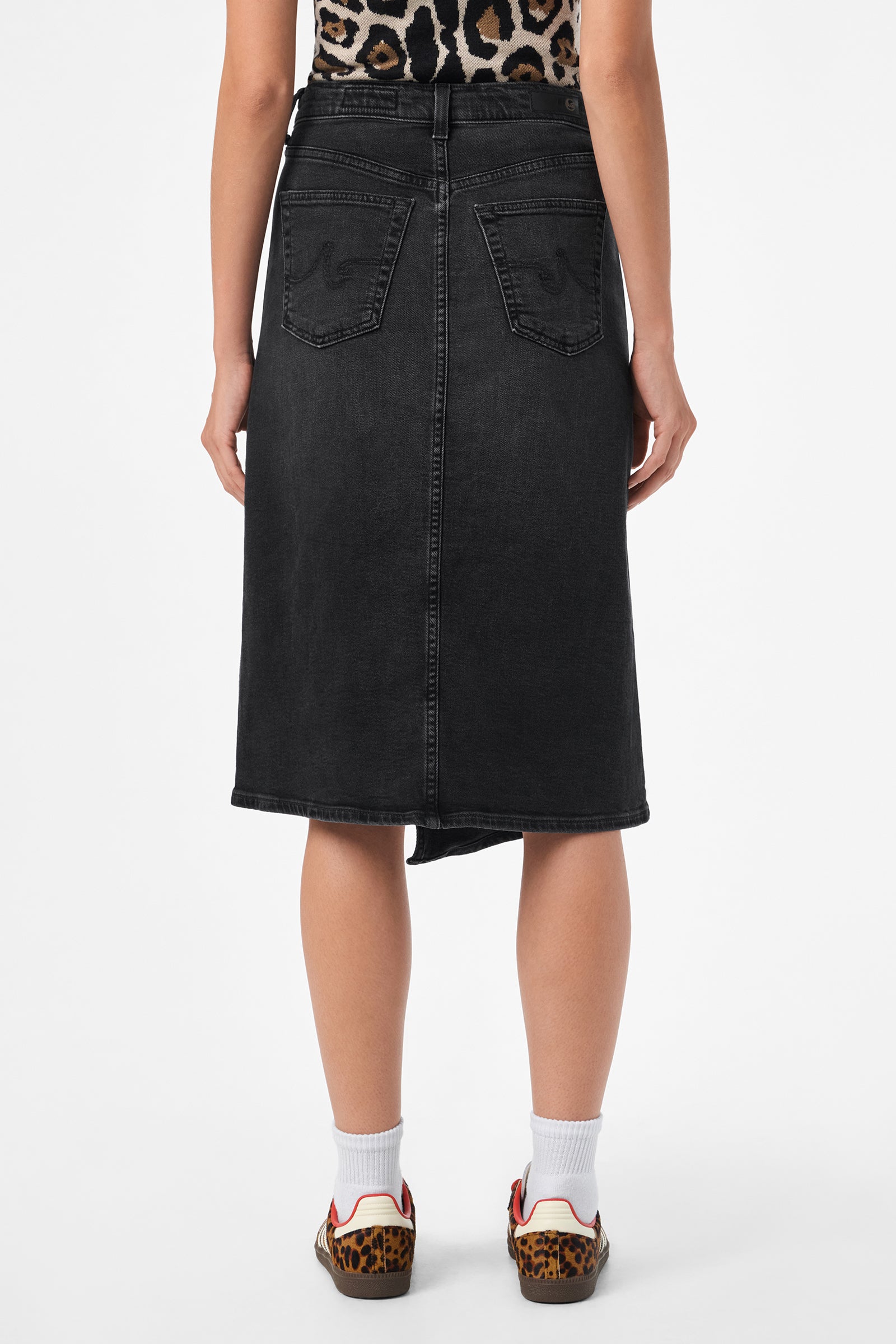 A person is shown from the back wearing the AG Bruna Skirt in black denim, paired with a leopard print top, white socks, and leopard print detail sneakers, against a plain white background.