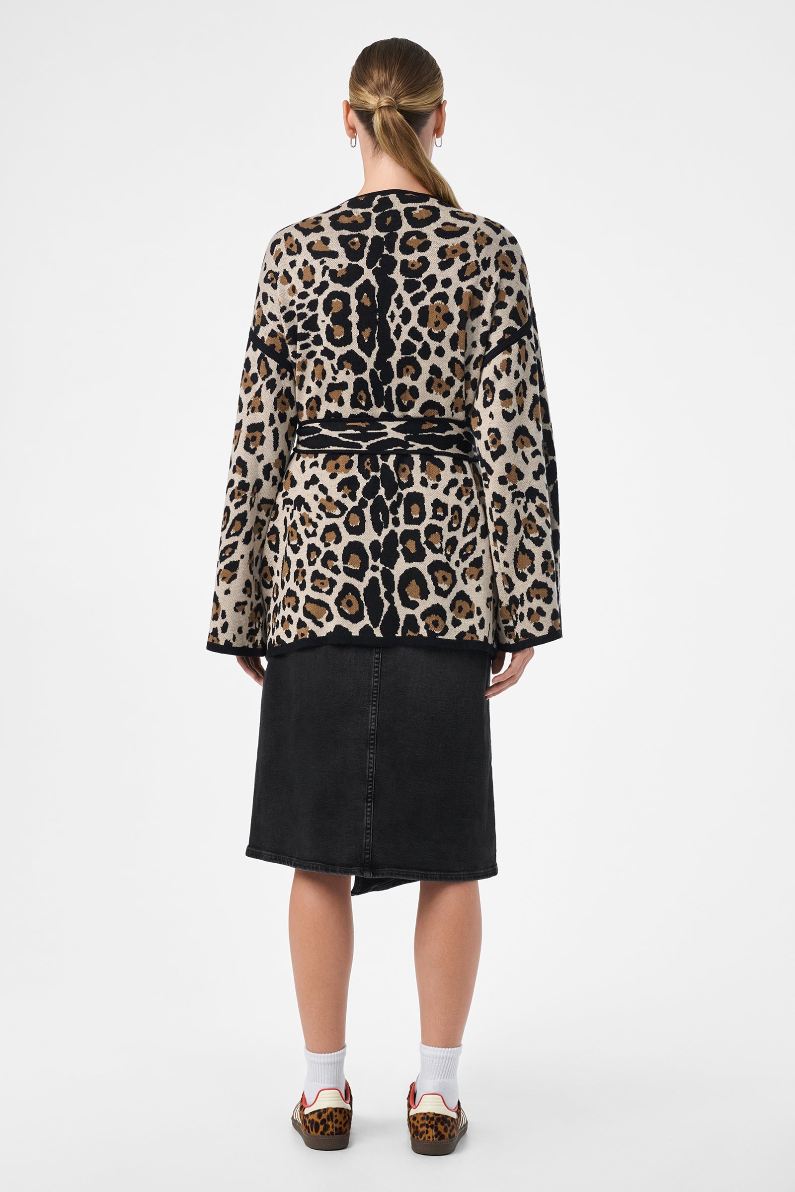 A person stands facing away, wearing the Wright Cardigan with a belt, a black knee-length skirt, white socks, leopard print loafers, and has their hair tied back in a low ponytail.