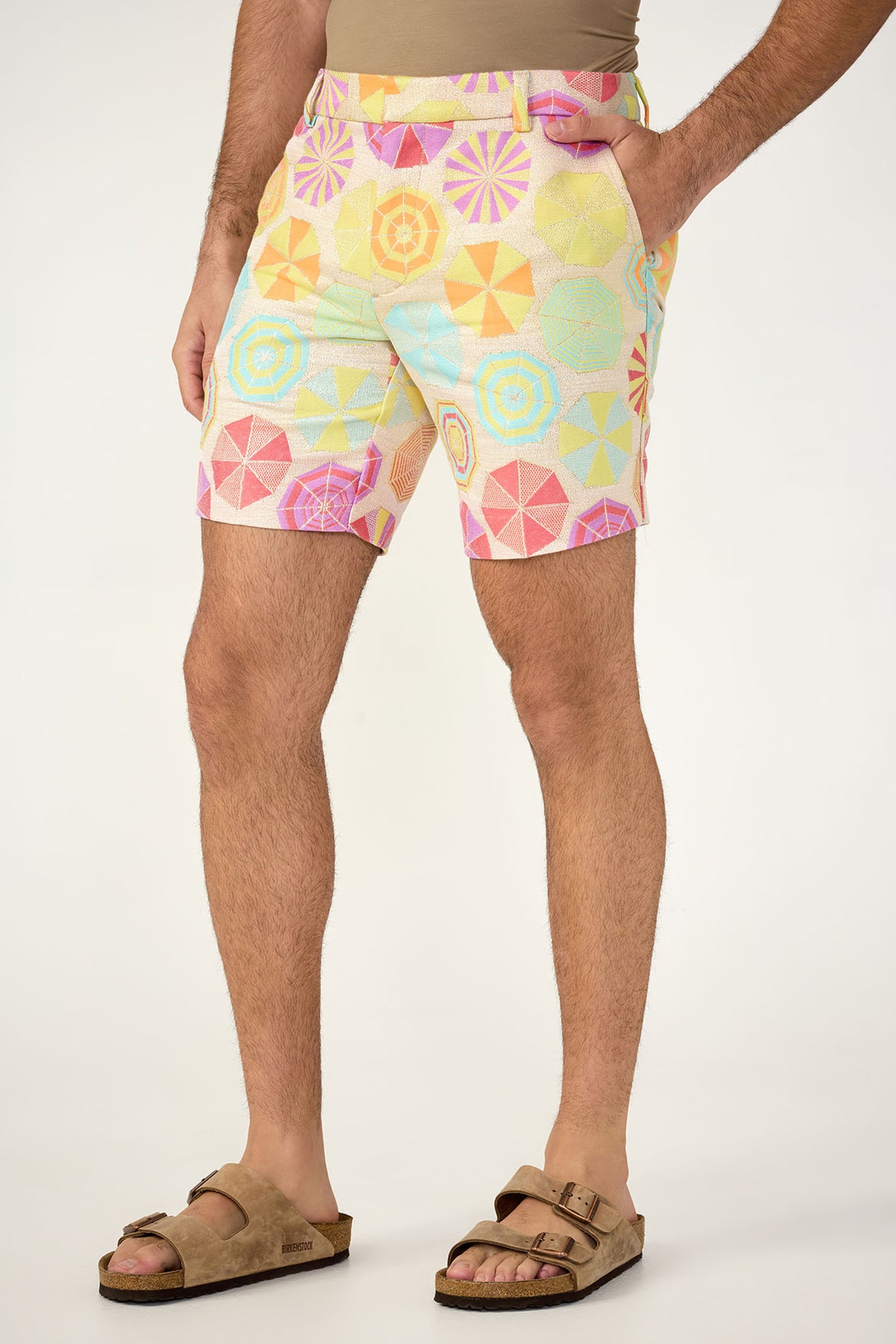 Wearing the Lawrence Short - Paraplue Jacquard, featuring a geometric umbrella pattern in pastel shades, paired with a beige shirt and brown sandals, standing before a simple light backdrop.