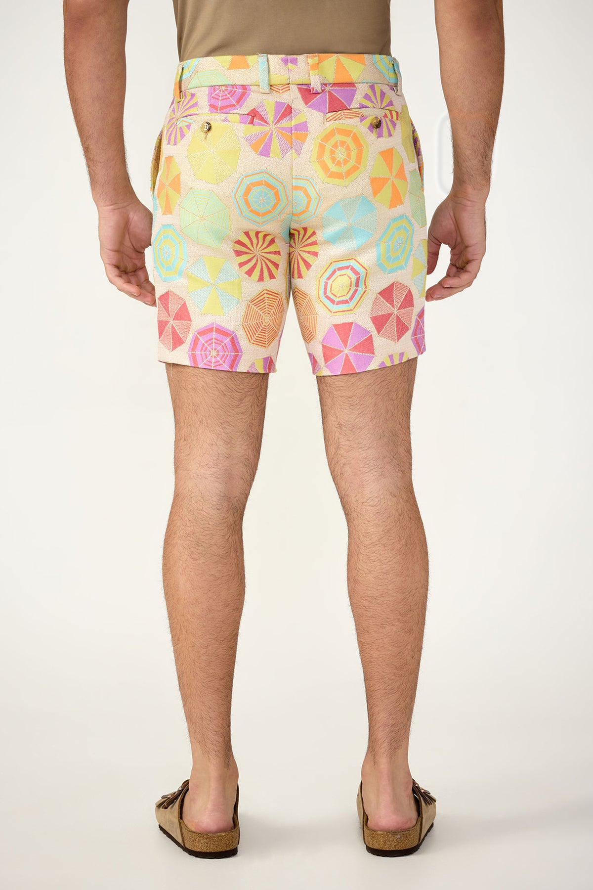 A man stands with his back to the camera, wearing tan sandals and Lawrence Short - Paraplue Jacquard featuring a pastel beach umbrella pattern. The background is plain and light.