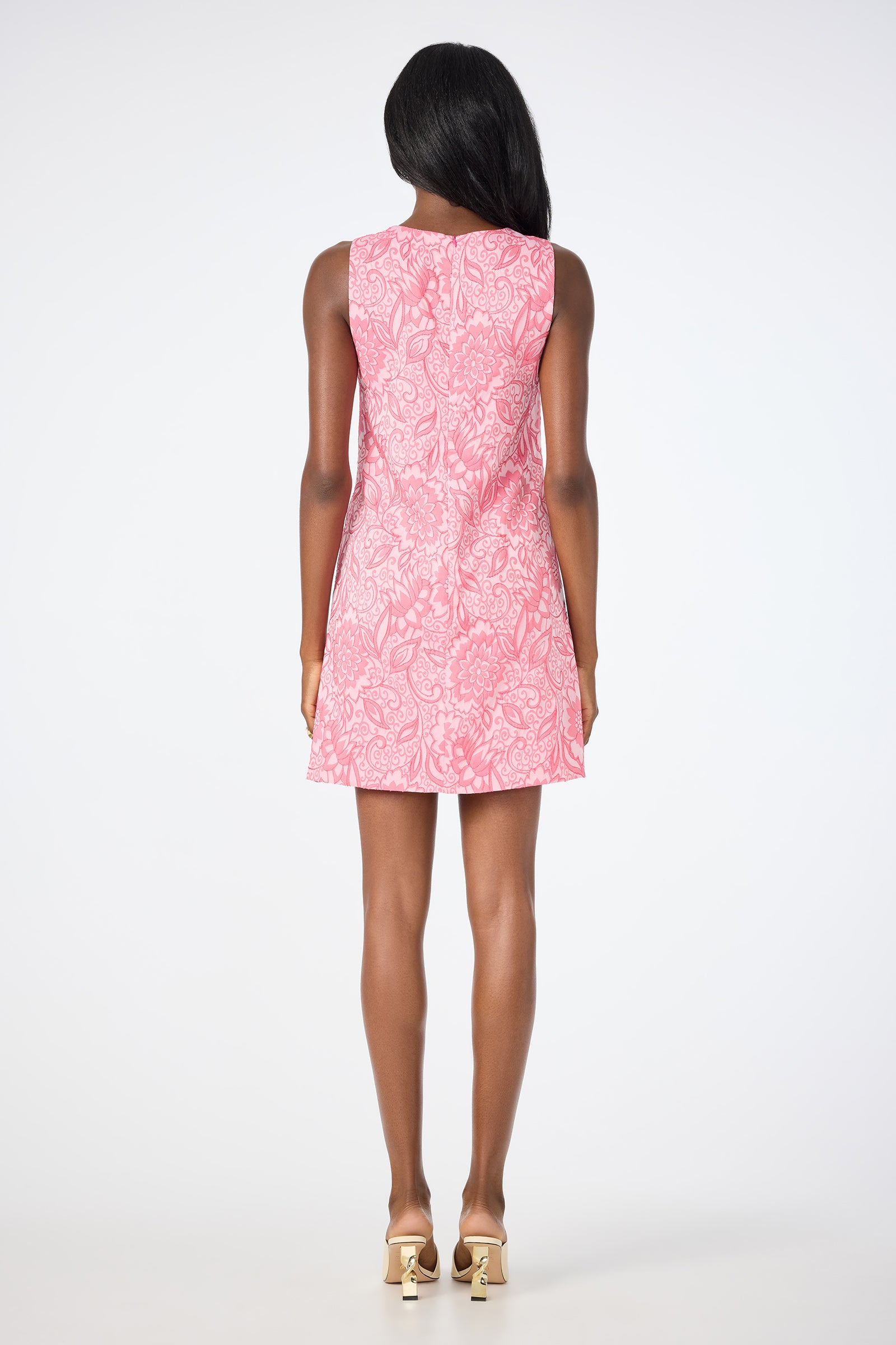 A woman faces away from the camera in the South Seas Sleeveless Dress - Jolie Jacquard, a pink floral dress, paired with gold high-heeled sandals against a white background.
