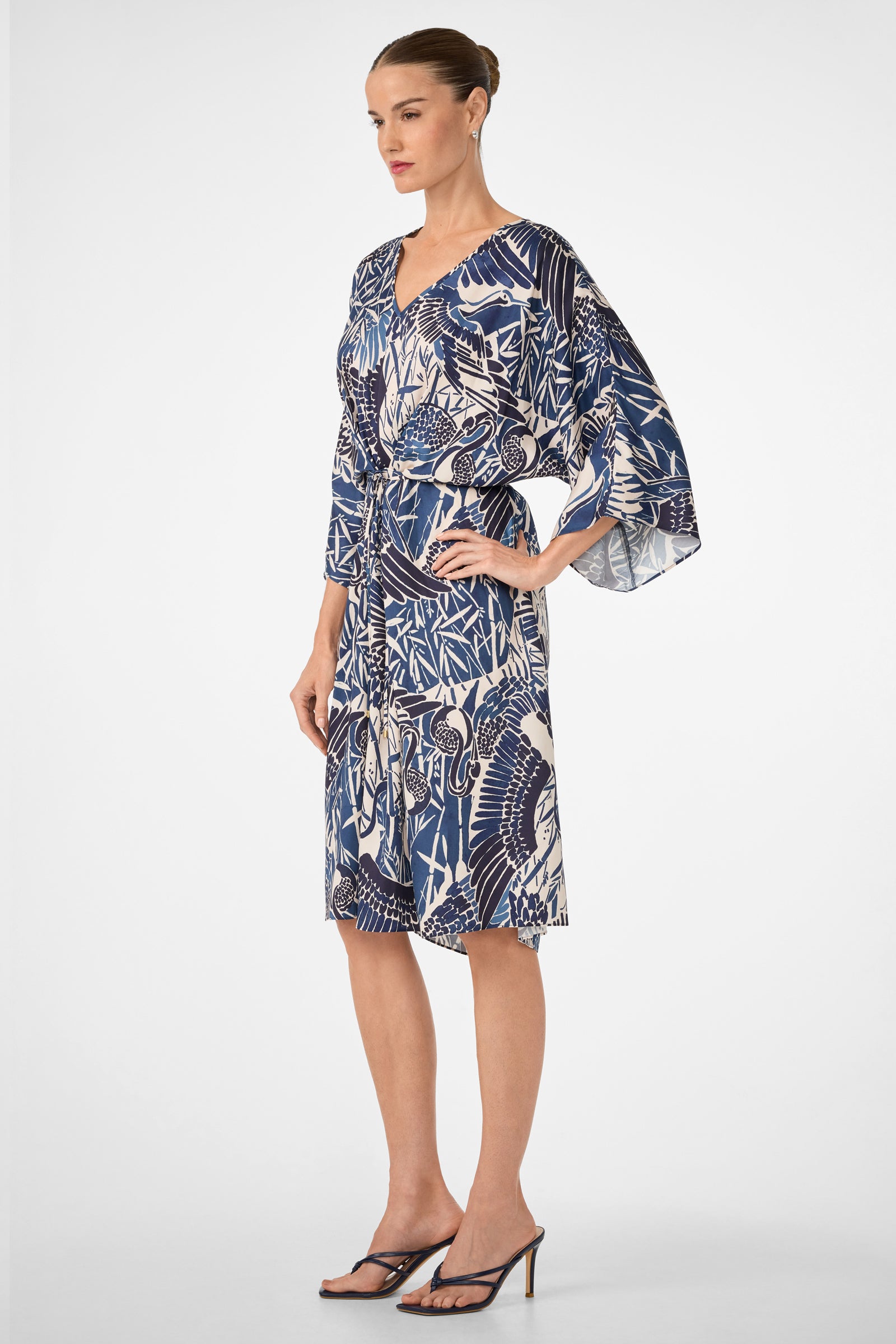 A woman in profile wears the Topspin Short-Sleeve Midi Dress - Tsuru, featuring blue and white patterns, a tied waist, and loose sleeves. She pairs it with black heeled sandals and has her hair in a neat bun against a plain white background.