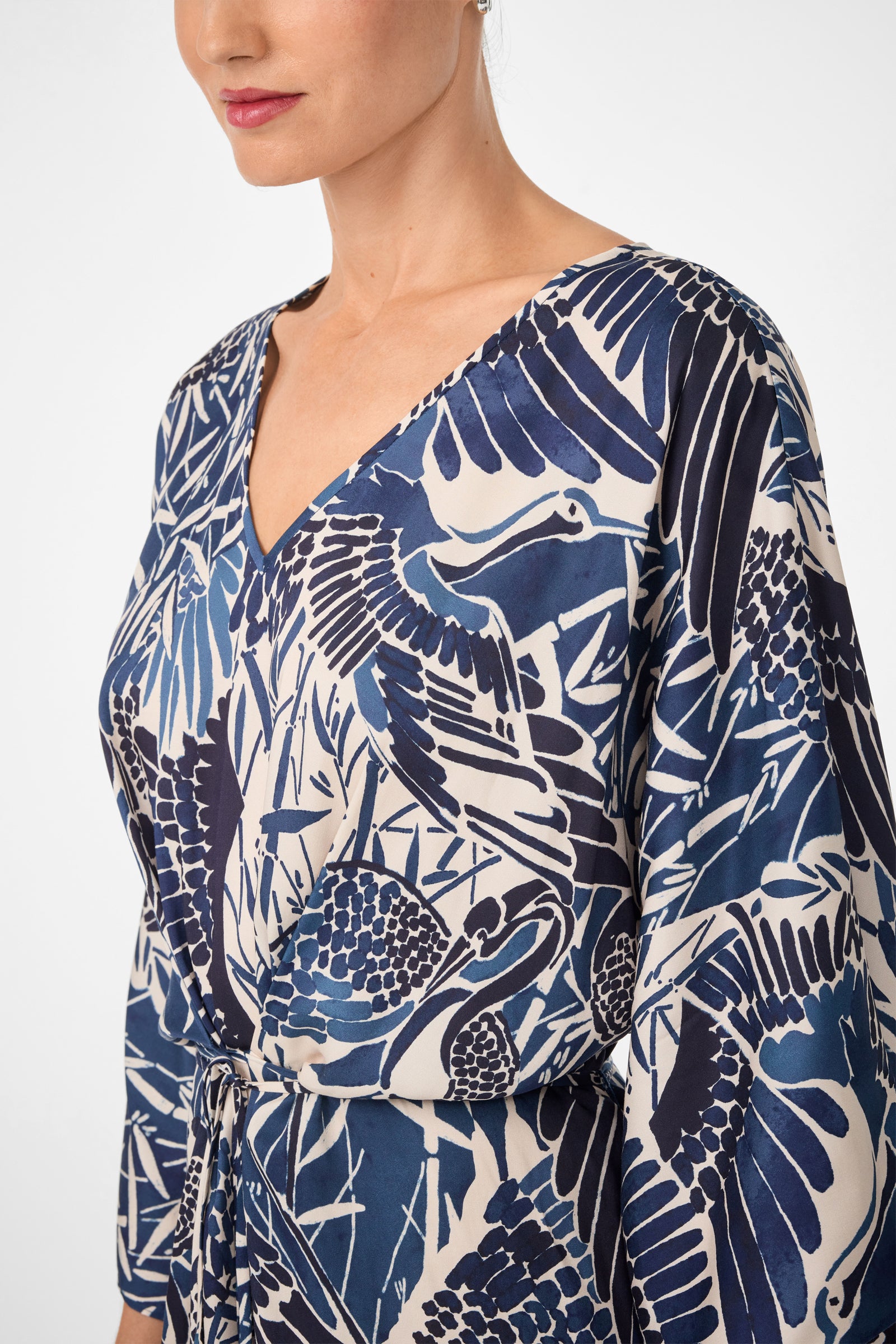A woman wears the Topspin Short-Sleeve Midi Dress - Tsuru, a blue and white V-neck dress with a nature-inspired pattern and drawstring waist. Photo shows her from shoulders to mid-torso against a plain background.