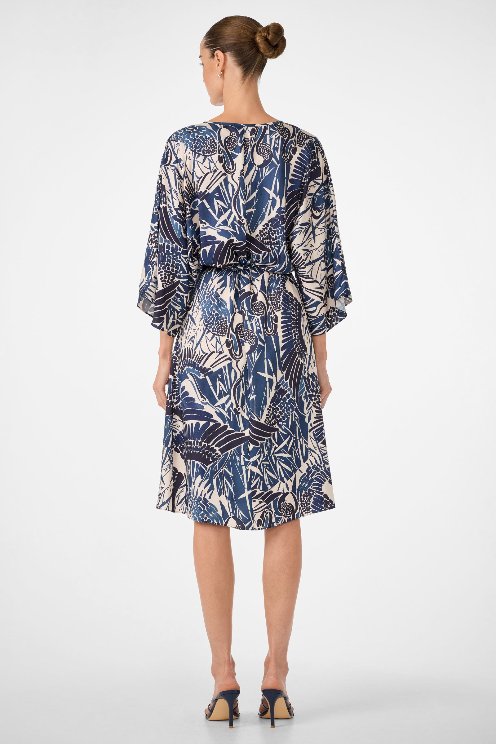 A woman faces away, wearing the Topspin Short-Sleeve Midi Dress - Tsuru, featuring blue and white patterns, loose sleeves, and a tied waist. Her hair is in a bun, paired with open-heeled sandals against a plain background.