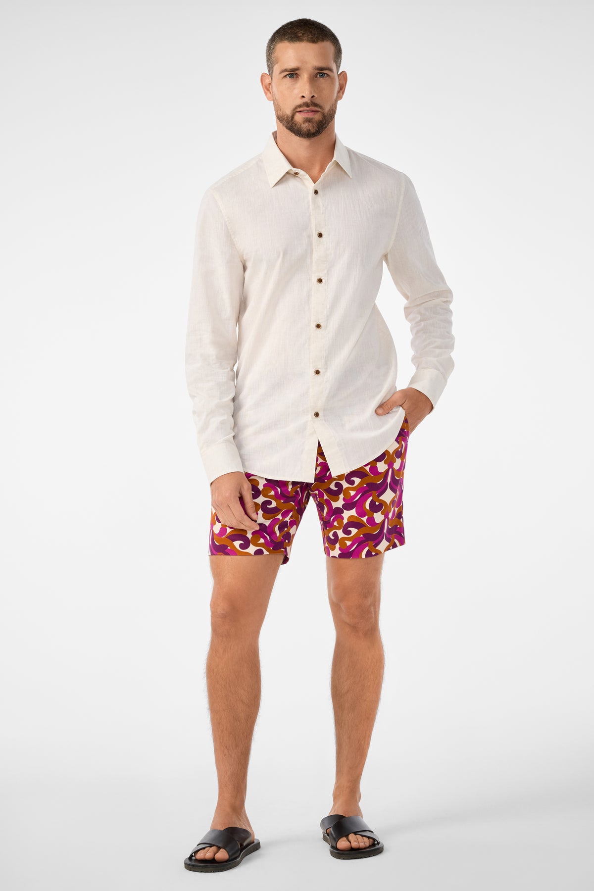 A man with short hair and a beard poses with one hand in his pocket, wearing the Lawrence Short—patterned magenta and yellow shorts—paired with a cream long-sleeve button-up shirt and black sandals against a plain background.