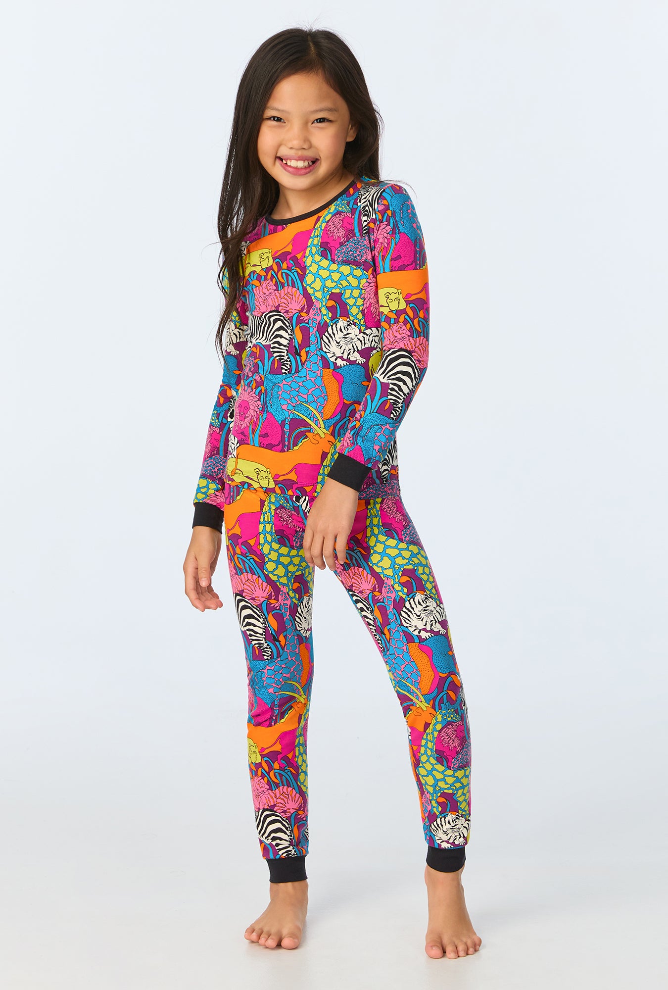 A smiling child with long dark hair models the Jungle Kids Long-Sleeve Long Pant Jersey PJ Set in Multi, featuring vibrant animal prints on white. She stands barefoot against a light backdrop, highlighting comfy and playful sleepwear.