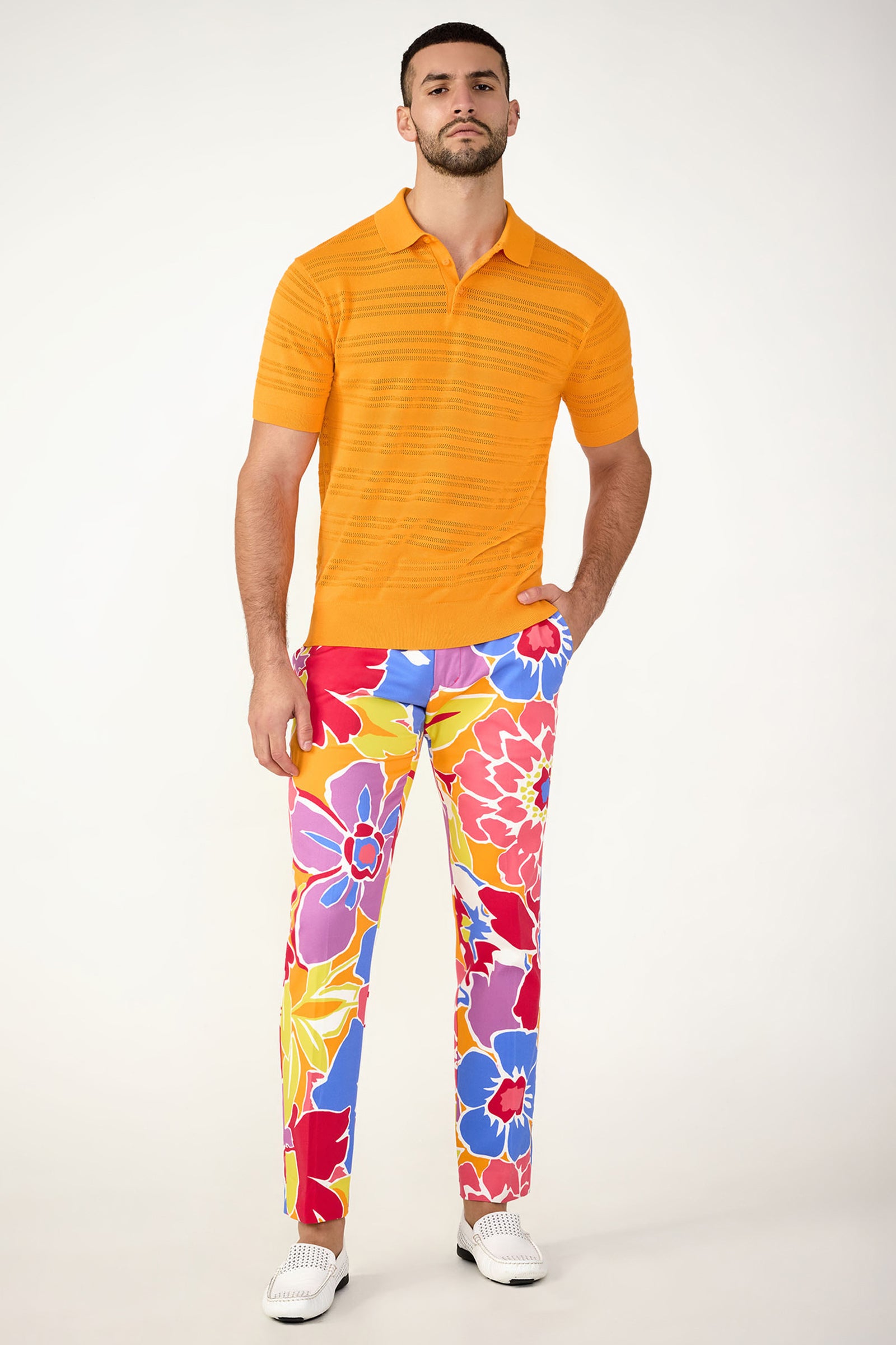 A man stands against a plain background wearing an orange polo, white shoes, and the Clyde Slim Trouser - Soie Paradis: brightly colored floral pants in red, yellow, blue, and purple. His hands are in his pockets and he has a neutral expression.
