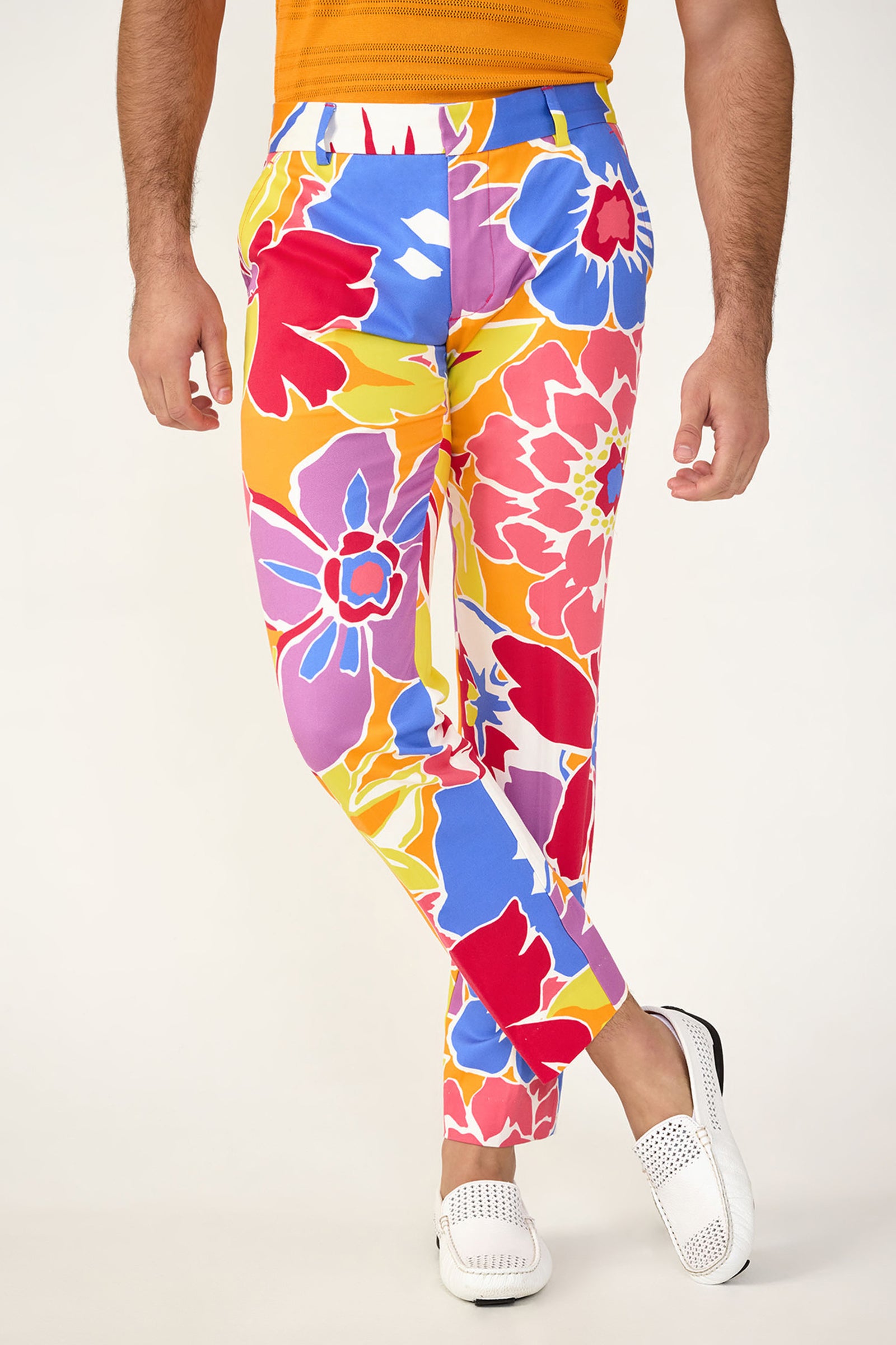 A person wears the Clyde Slim Trouser in Soie Paradis, featuring a bright floral pattern, paired with an orange shirt and white loafers, standing against a plain light background.