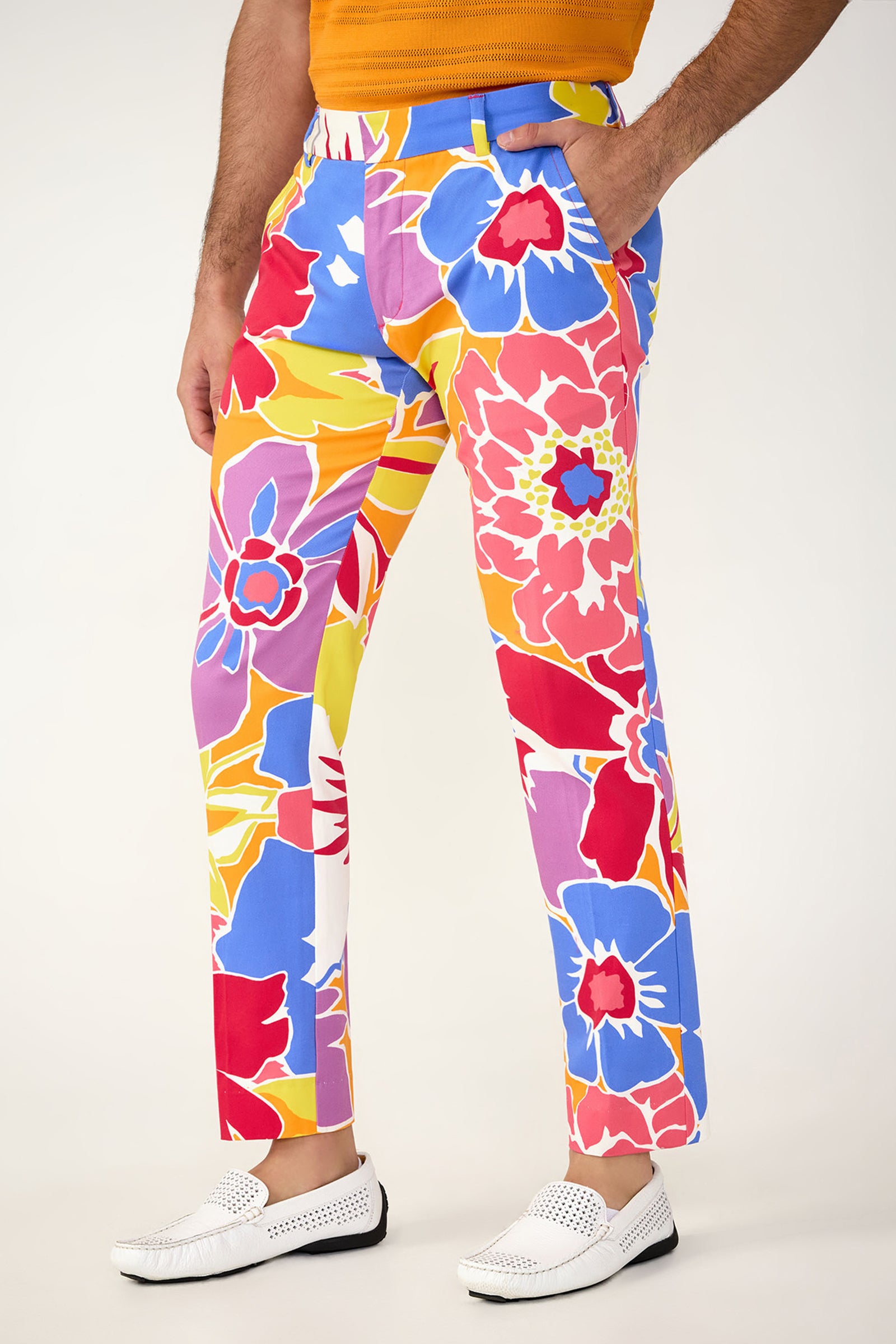 A person wears the Clyde Slim Trouser - Soie Paradis, featuring a bold, oversized floral pattern in red, blue, yellow, and pink. Paired with a bright orange top and white loafers. Only the lower half of the body is visible.