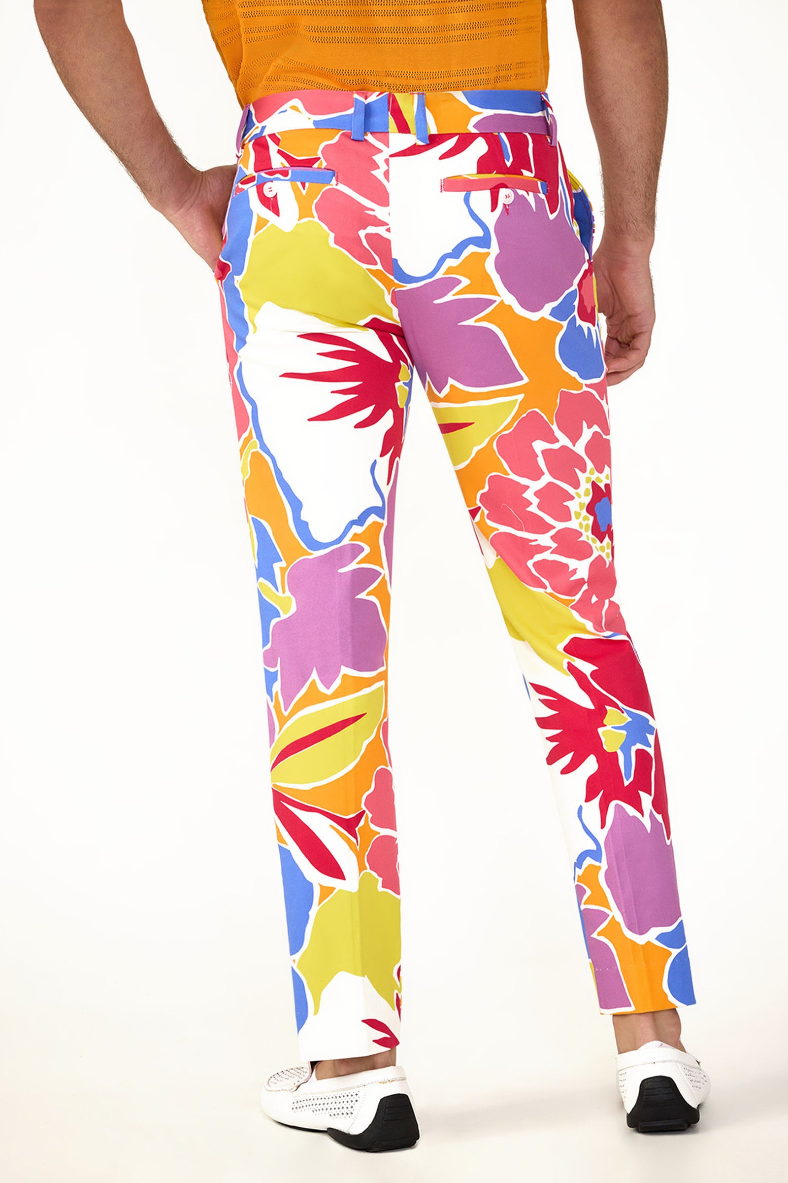 A person wearing the Clyde Slim Trouser - Soie Paradis, featuring a vibrant floral pattern in red, orange, yellow, pink, blue, and white, paired with a tucked-in orange shirt and white shoes, shown from the back.