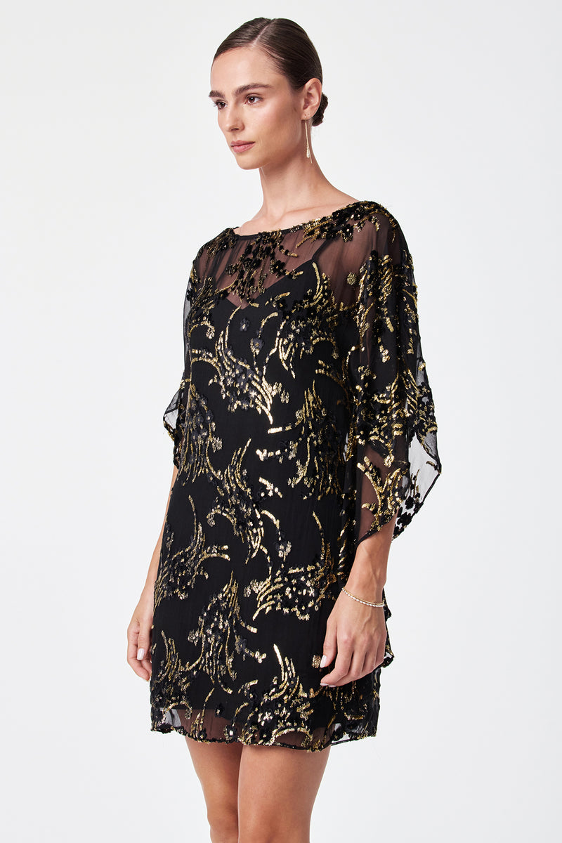 A woman stands against a white background wearing the Anissa Metallic Burnout Mini Caftan Dress, featuring sheer loose sleeves and a gold sequin pattern. Her hair is pulled back, and she accessorizes with a bracelet and small earrings.
