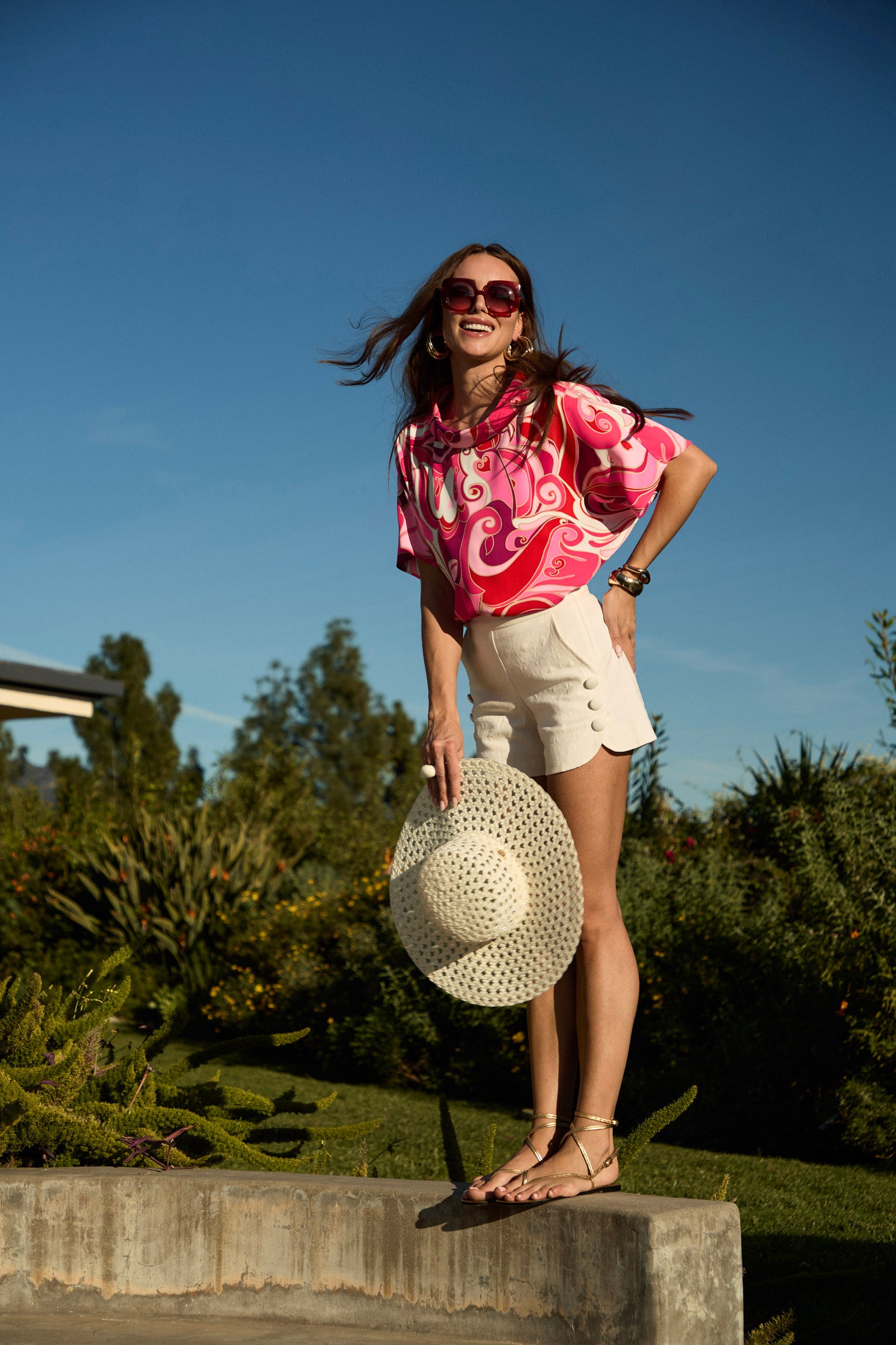 A woman in the Honeysuckle Boatneck Top, white shorts, and sunglasses stands outdoors on a low wall holding a white sunhat, smiling with greenery and a clear blue sky behind her.