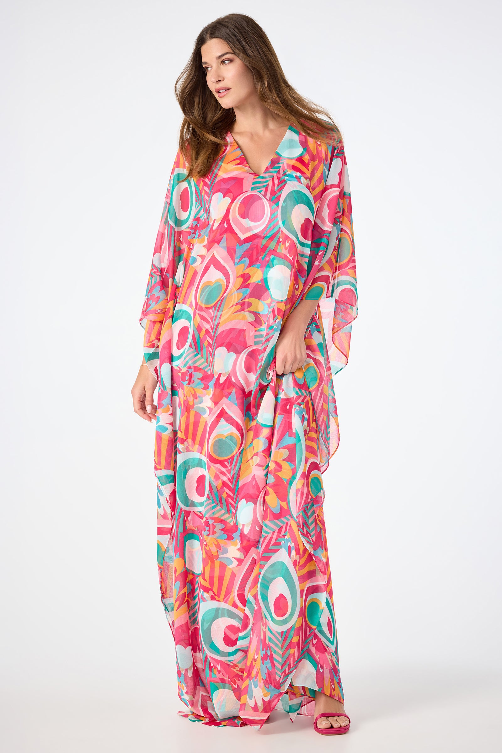 A woman wearing the Salon Maxi Caftan Dress - Dramont, featuring a bold abstract pattern in pink, green, orange, and white, stands against a plain white background.