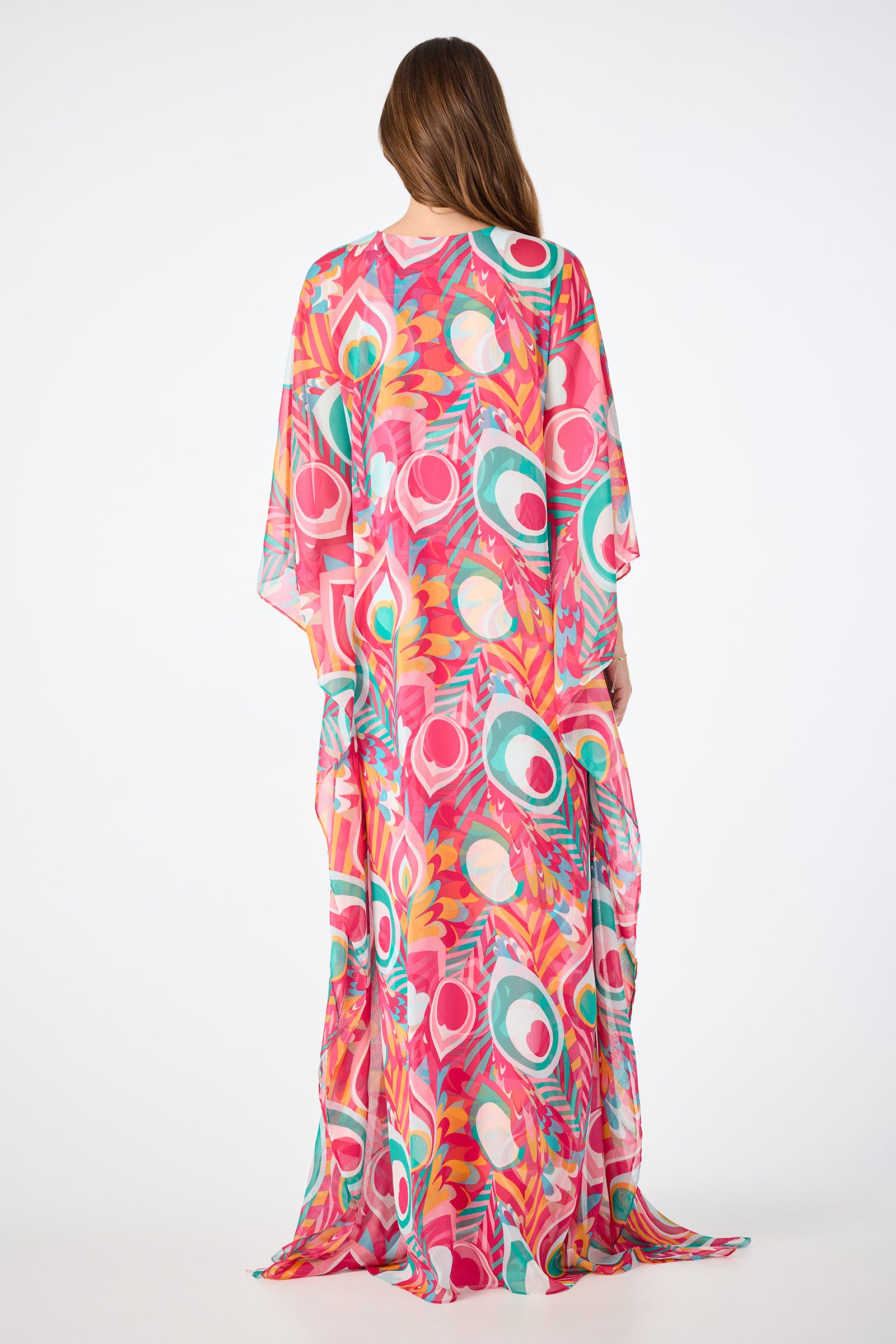 A woman with long brown hair, seen from behind, wears the Salon Maxi Caftan Dress – Dramont, featuring a flowing silhouette and vibrant peacock feather and floral patterns in pink, teal, green, and orange against a plain white background.