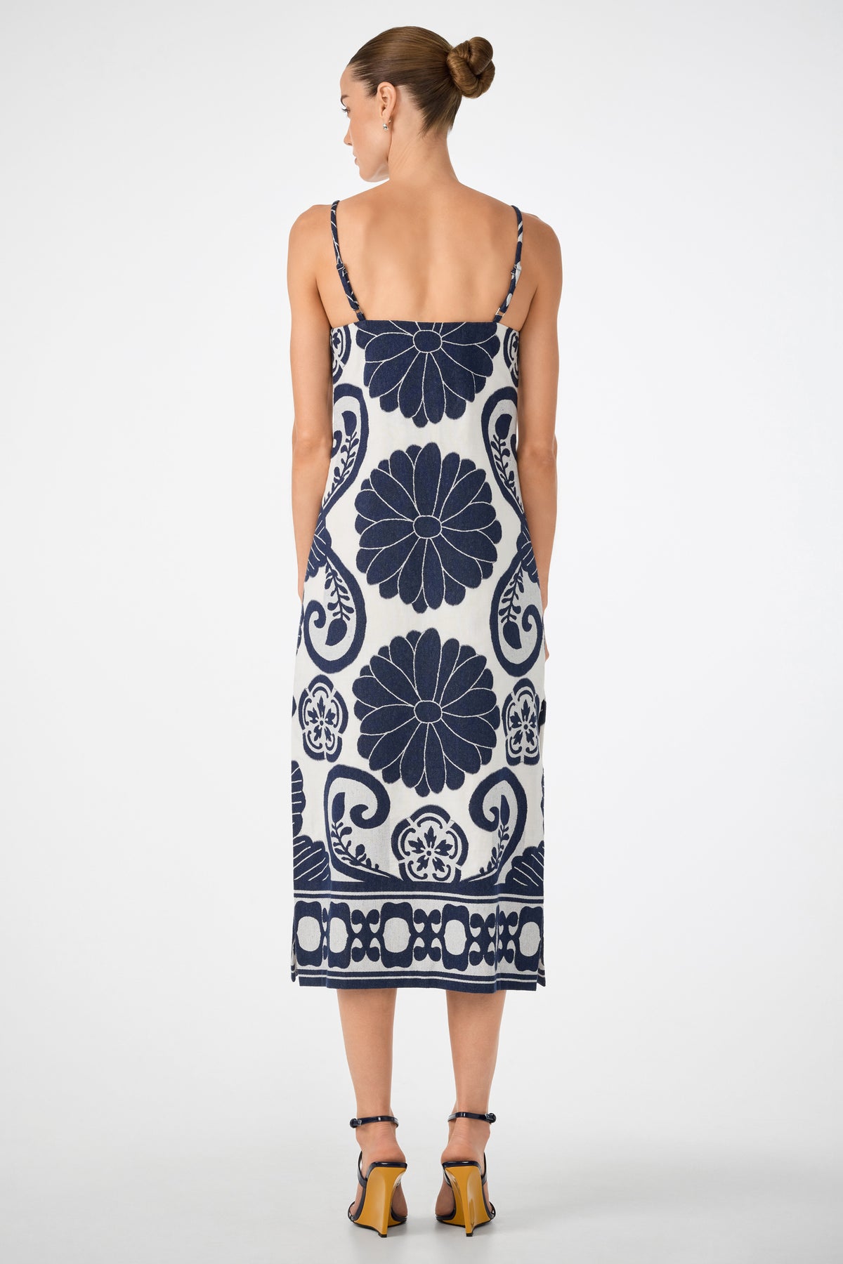 A woman with her hair in a bun is seen from behind, wearing the Marita 3 Sleeveless Dress—a white midi dress with thin straps and navy blue floral and geometric patterns—paired with strappy high-heeled sandals.