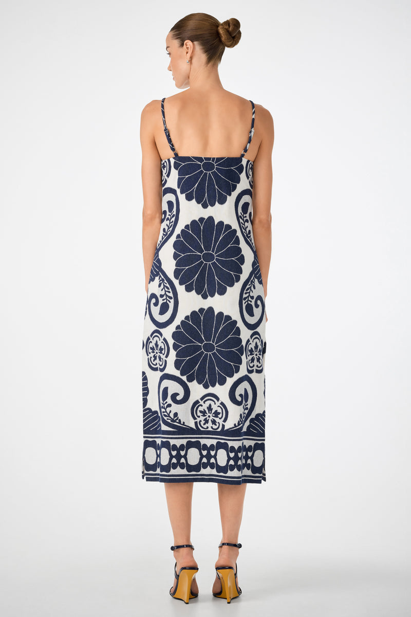 A woman with her hair in a bun is seen from behind, wearing the Marita 3 Sleeveless Dress—a white midi dress with thin straps and navy blue floral and geometric patterns—paired with strappy high-heeled sandals.