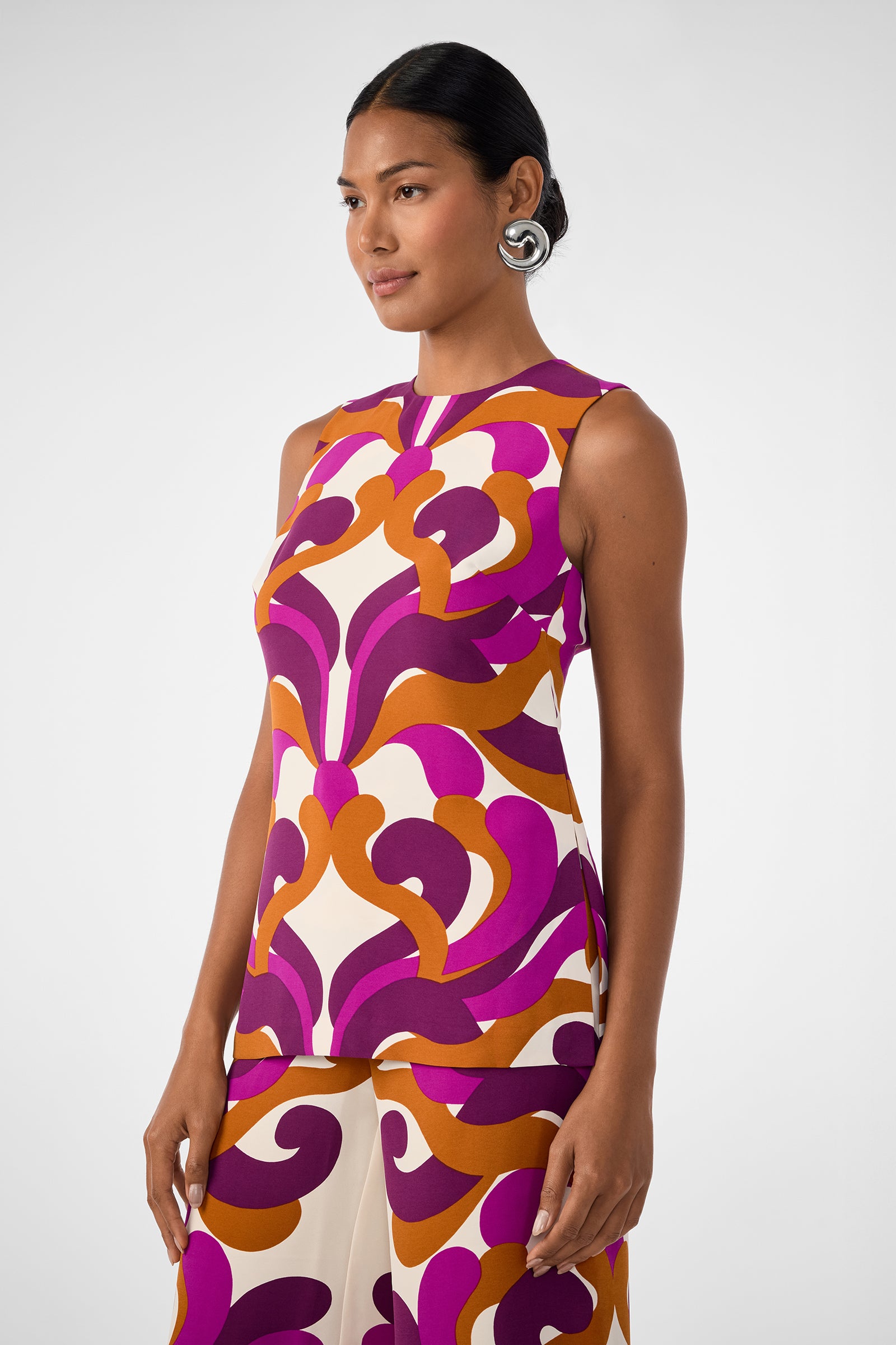 A woman with dark hair pulled back wears the Haruto Top, a sleeveless piece with a bold purple, orange, and cream pattern. She stands against a plain light background, looking slightly to the side.